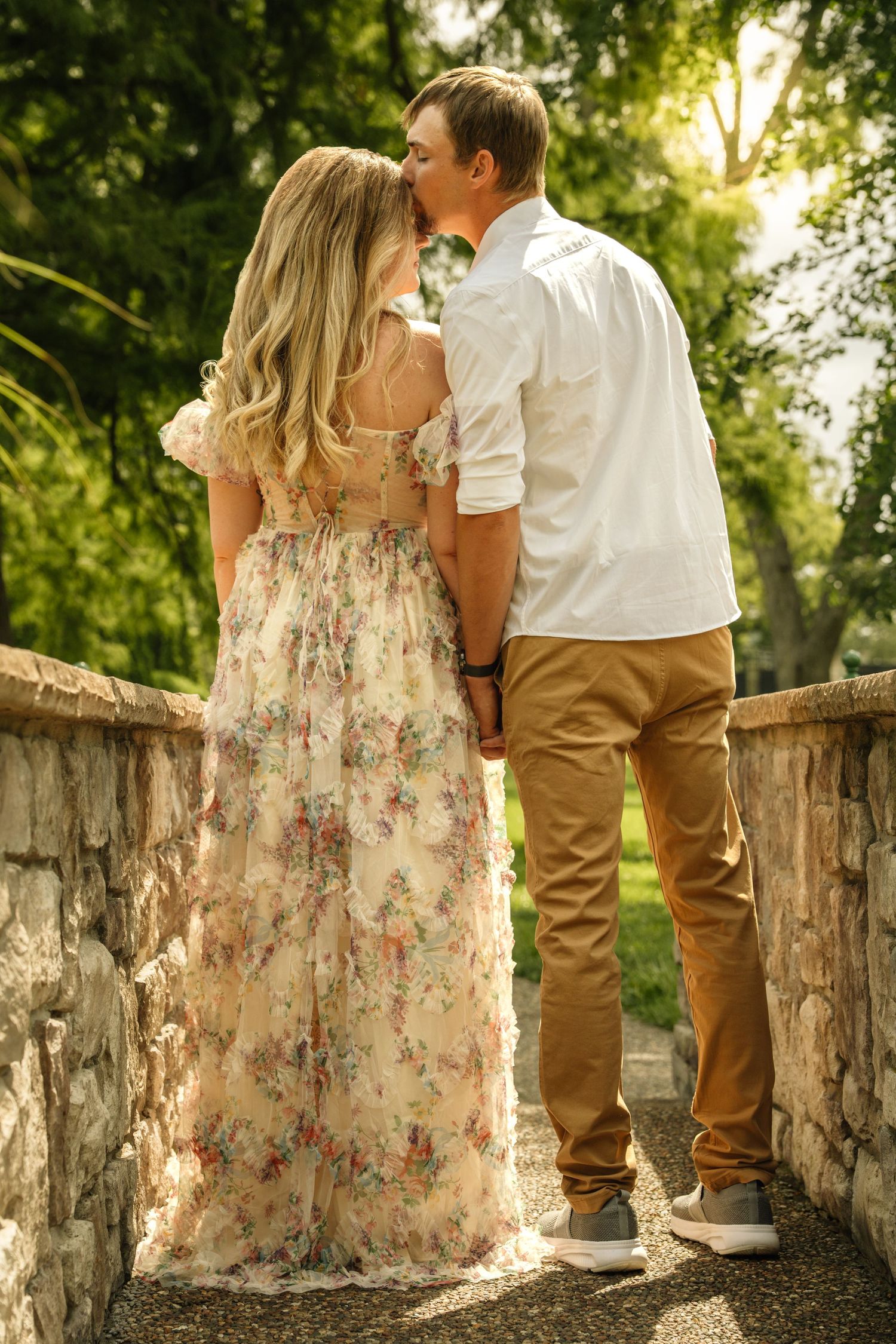 Couple sharing a kiss while holding hands on a stone bridge path in a sunlit park.