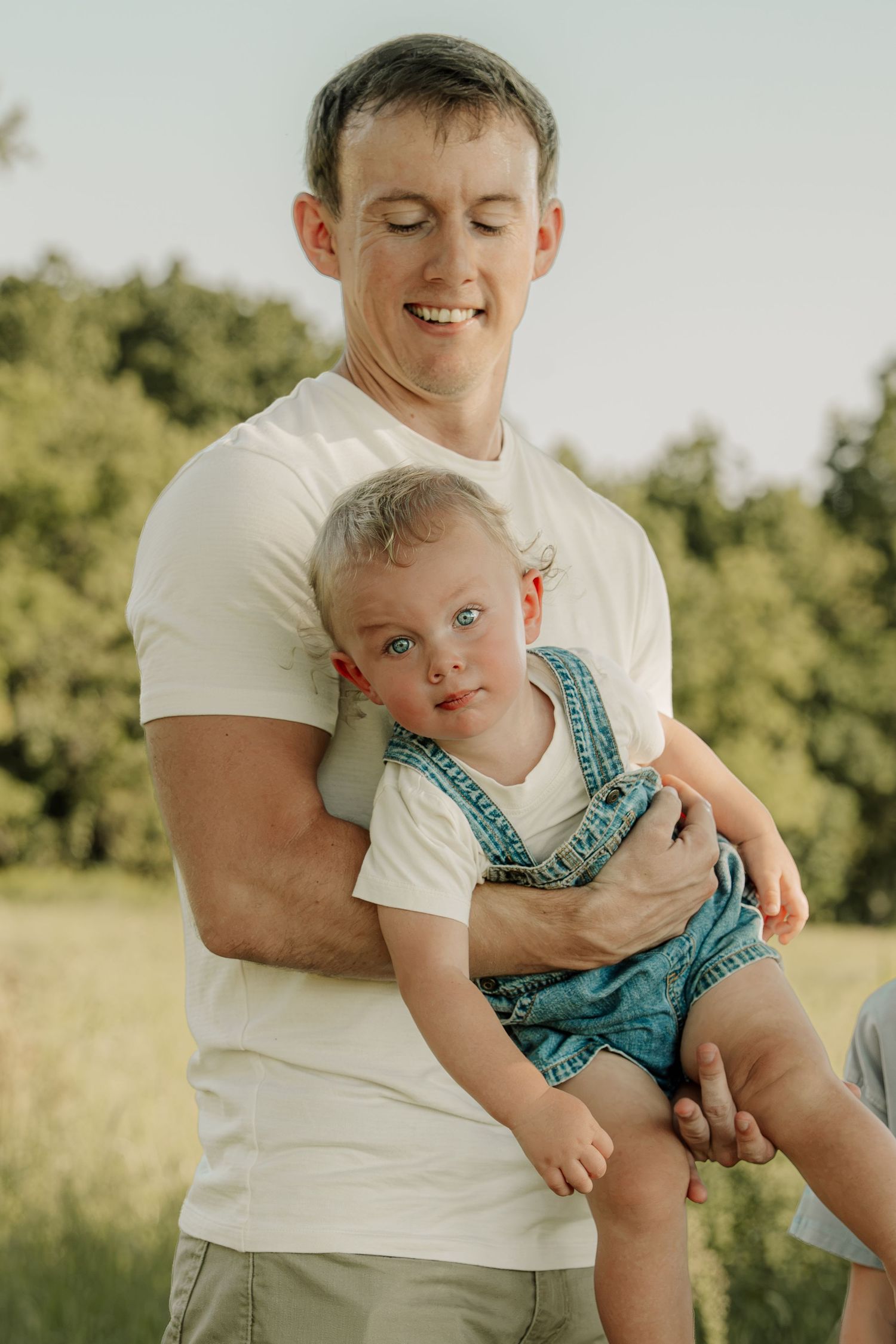 Smiling man in white t-shirt holding a baby in denim overalls outdoors in a green field.