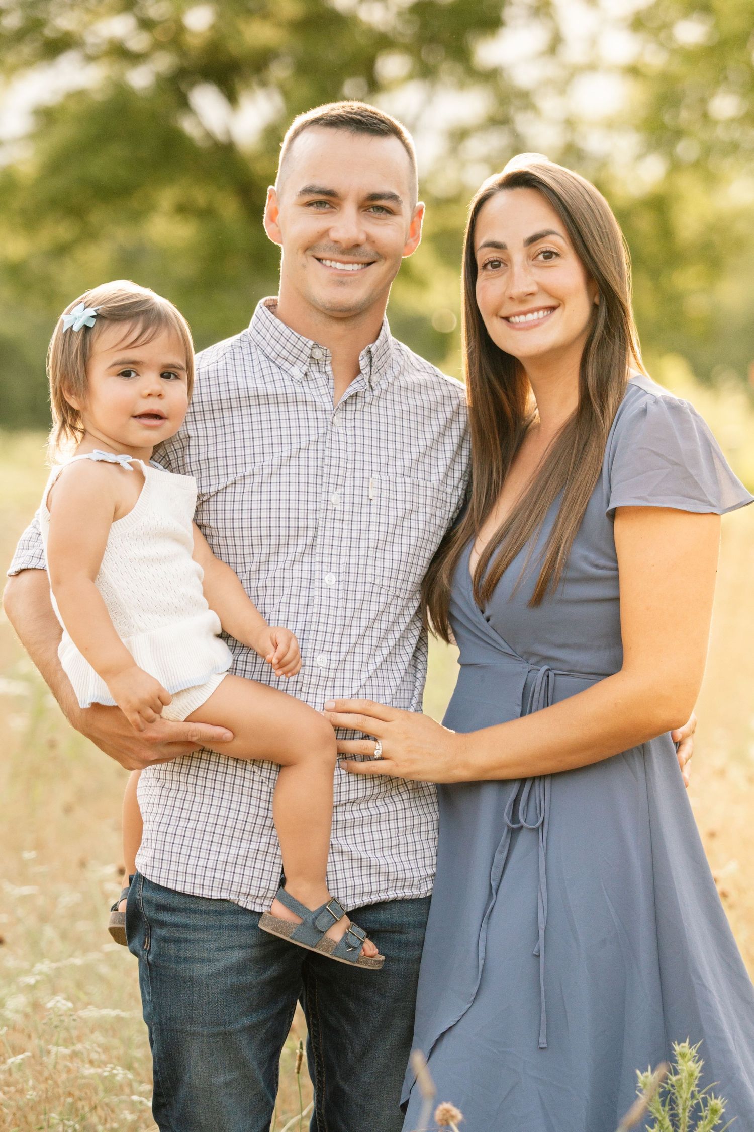 Happy young family with baby girl posing in a sunlit field, father holding child in blue dress.