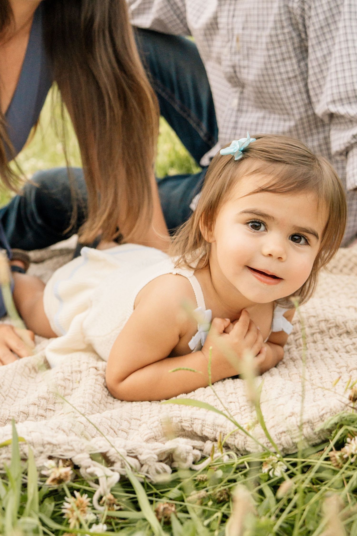 Adorable toddler girl lying on a blanket smiling at camera with parents nearby.