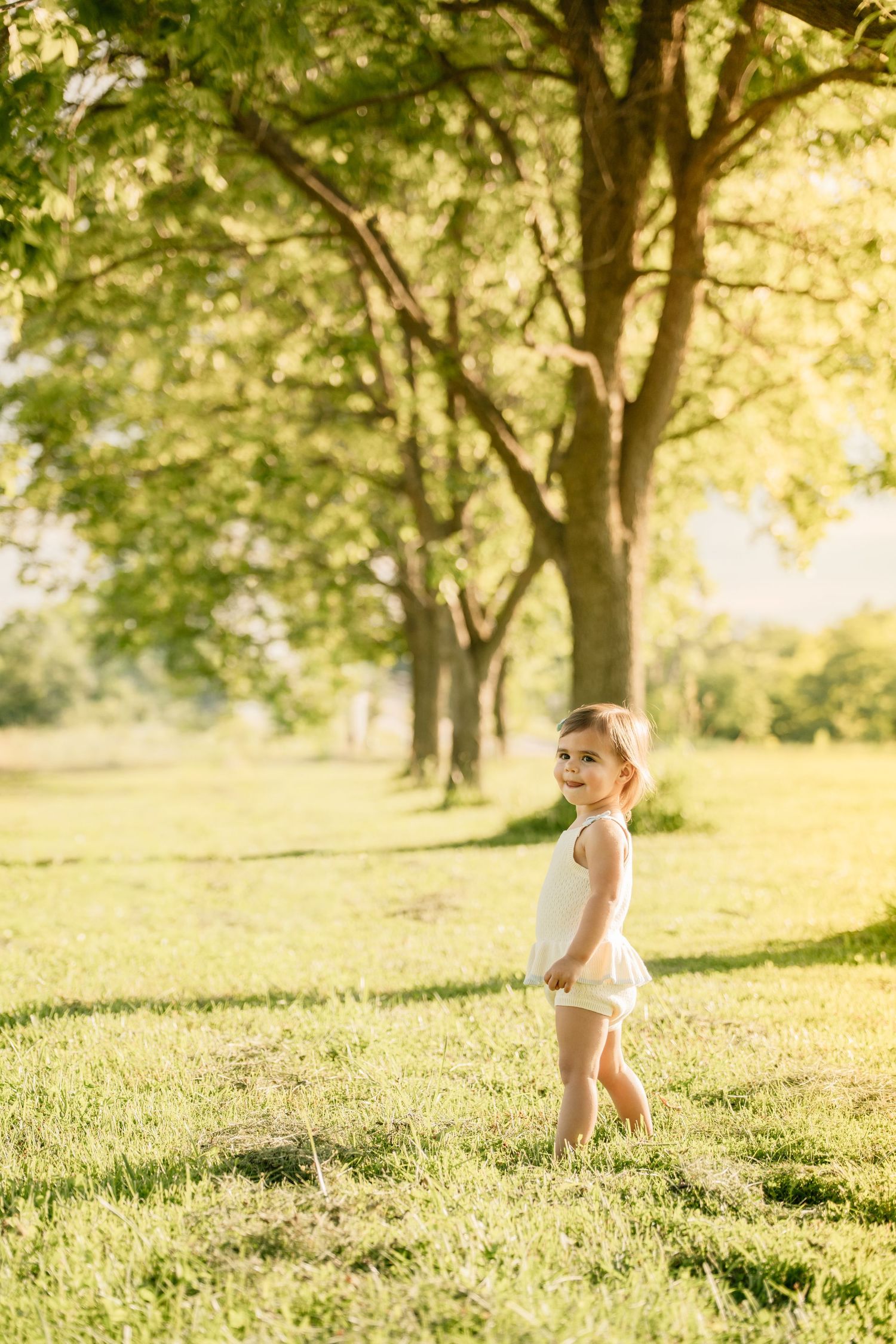 Toddler girl in white outfit standing on green grass in a sunny park with large trees.