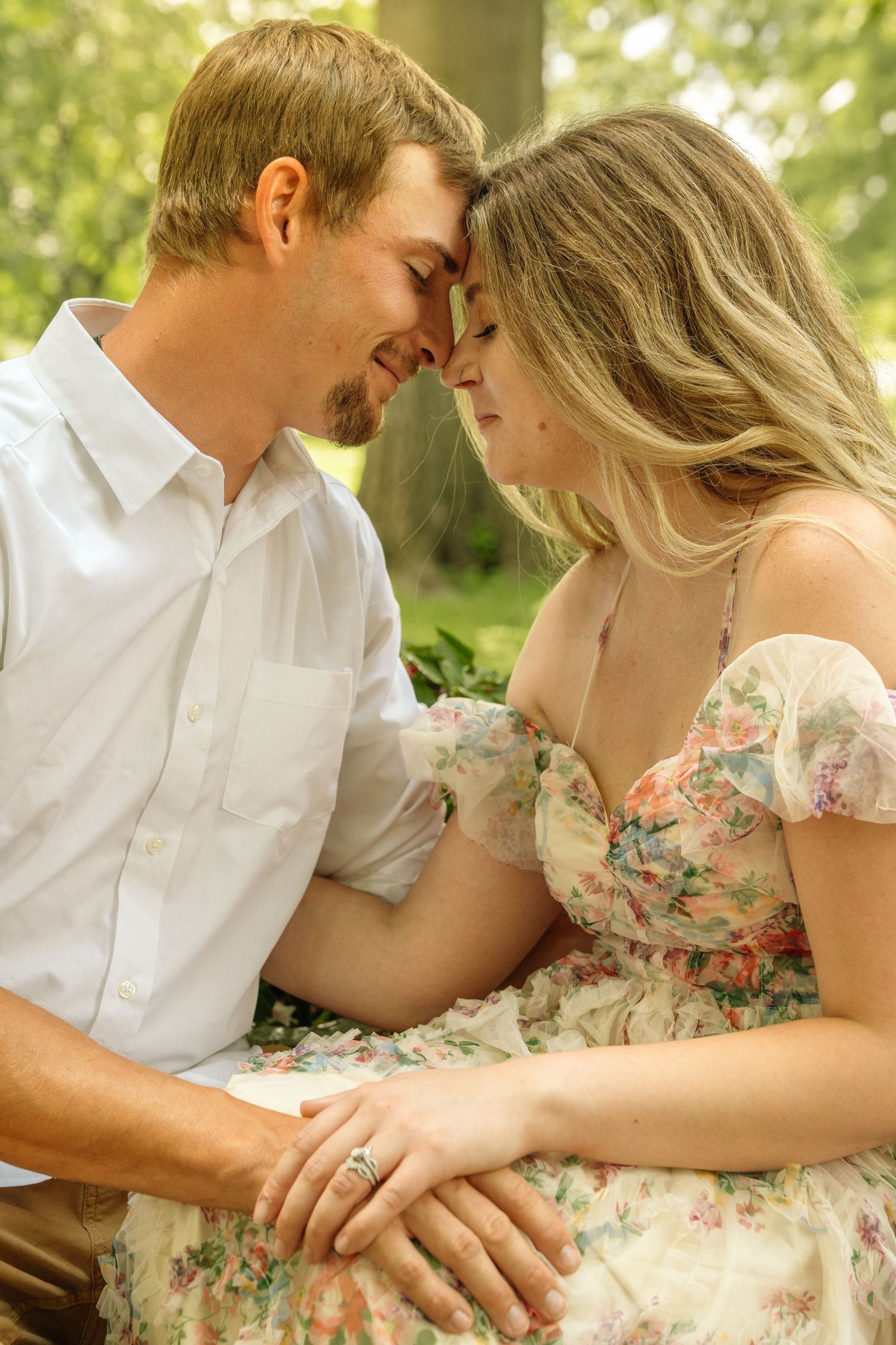Couple sitting close together touching foreheads tenderly with hands intertwined in a sunlit park.
