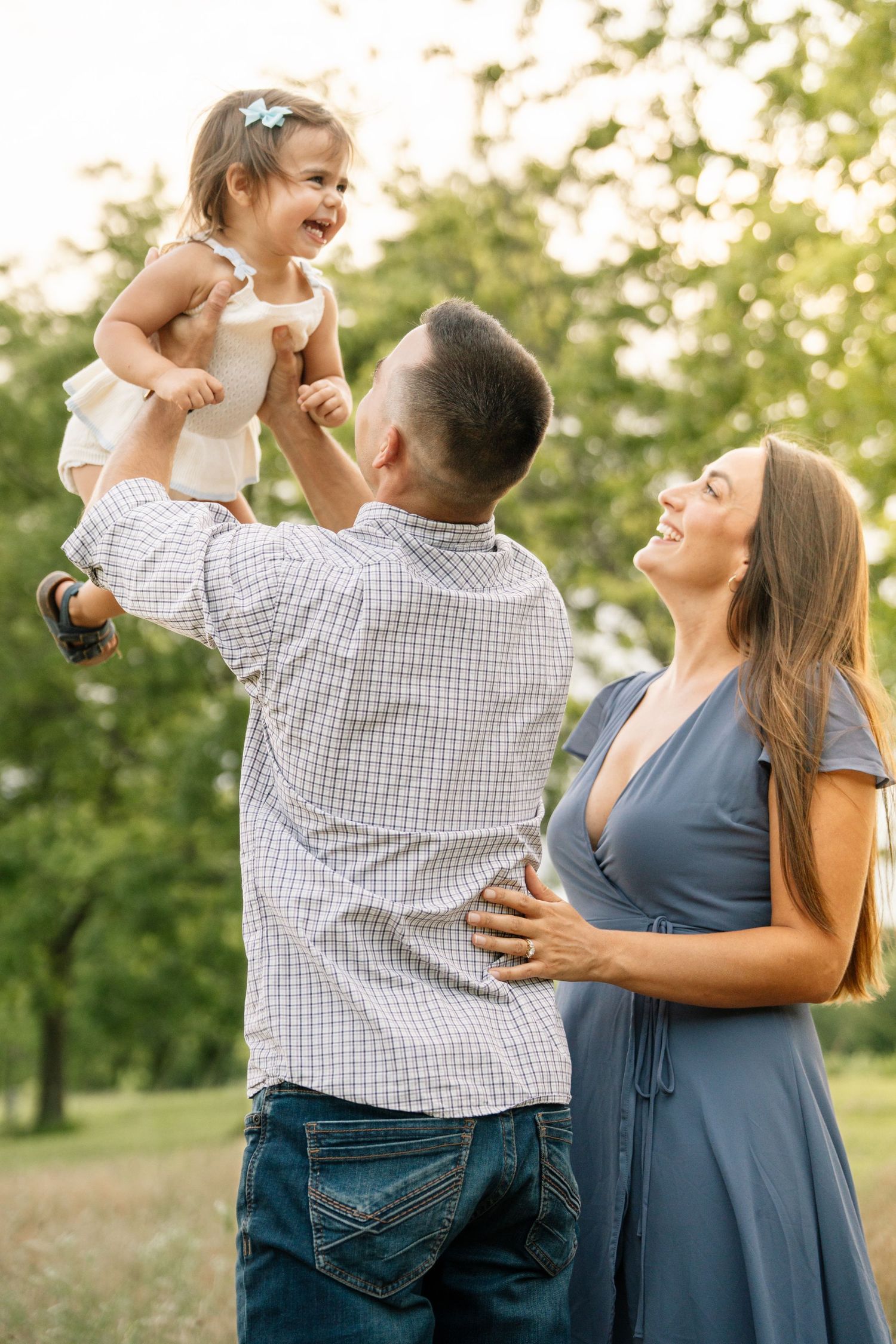 Happy family in a park, father lifting laughing toddler girl while pregnant mother smiles in a blue dress.