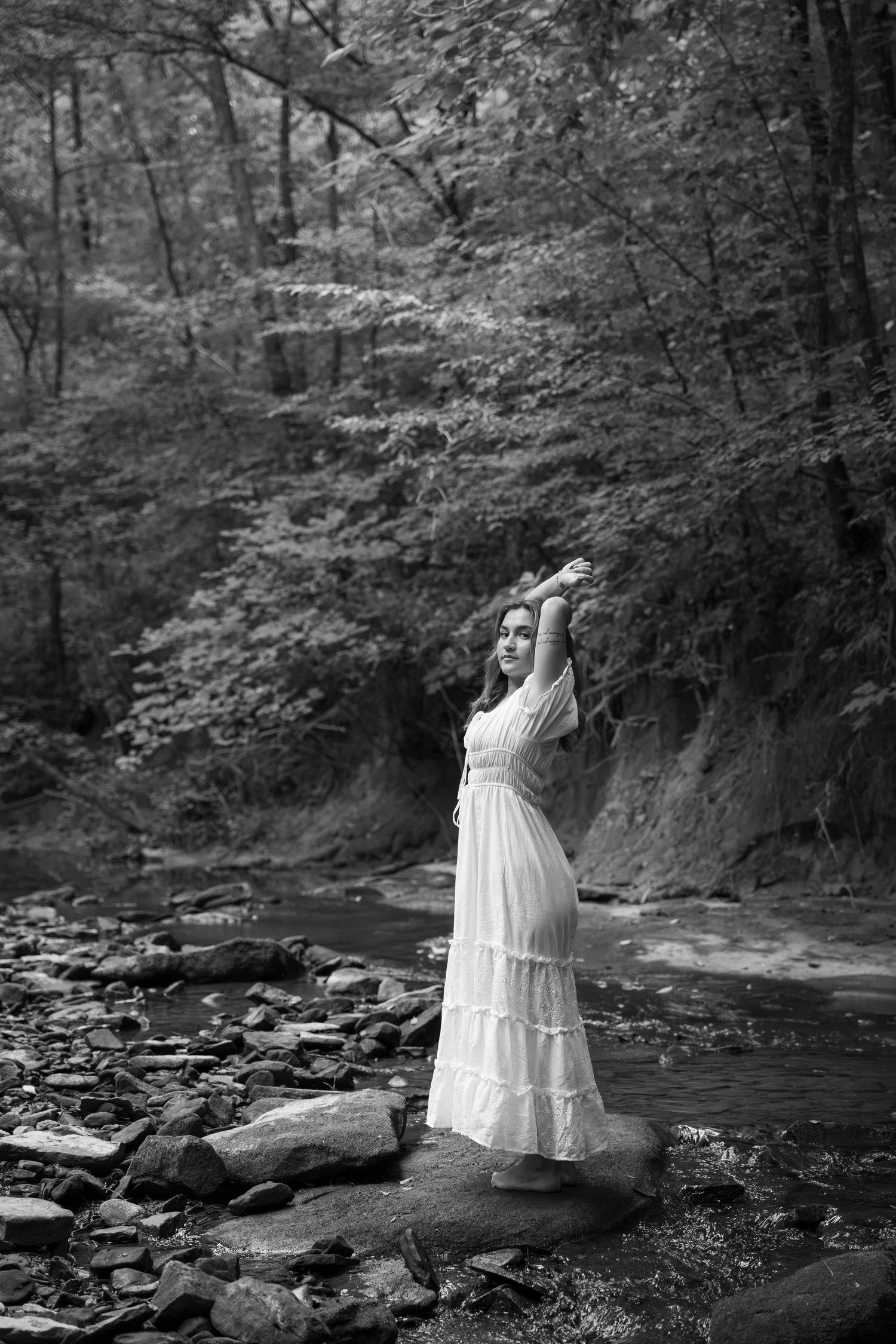 Black and white photo of a woman in white dress posing on rocks by a forest creek.