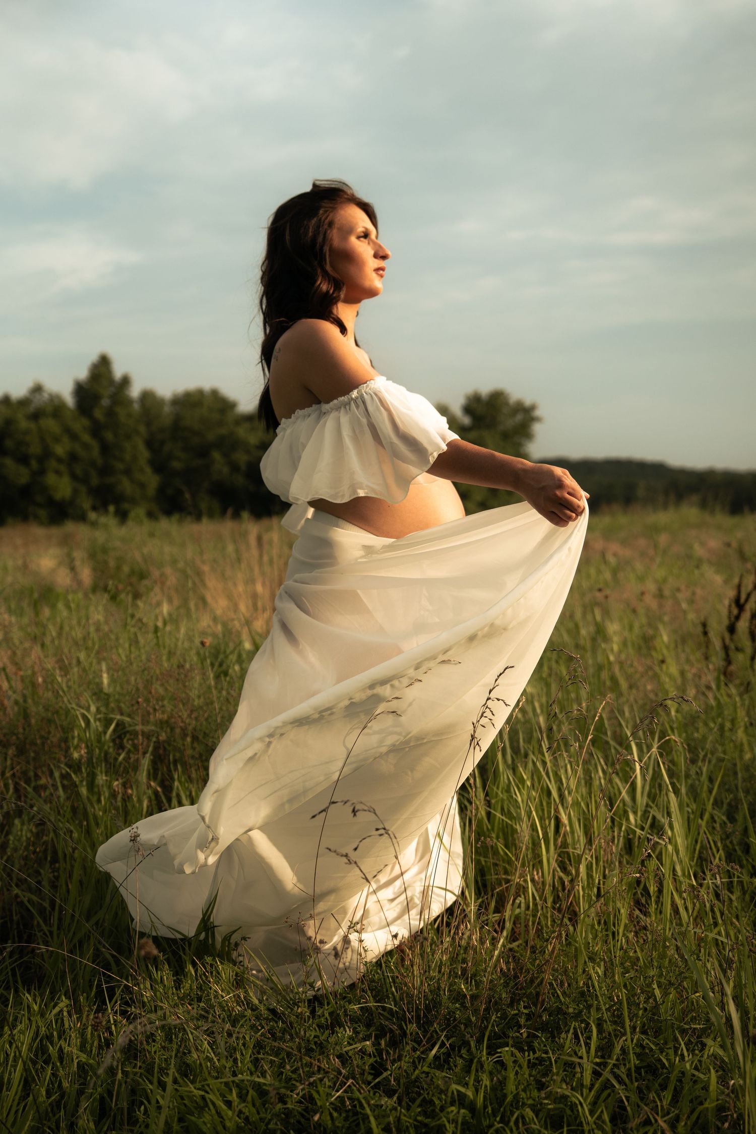 Pregnant woman in flowing white dress lifting her skirt in a golden sunset meadow.