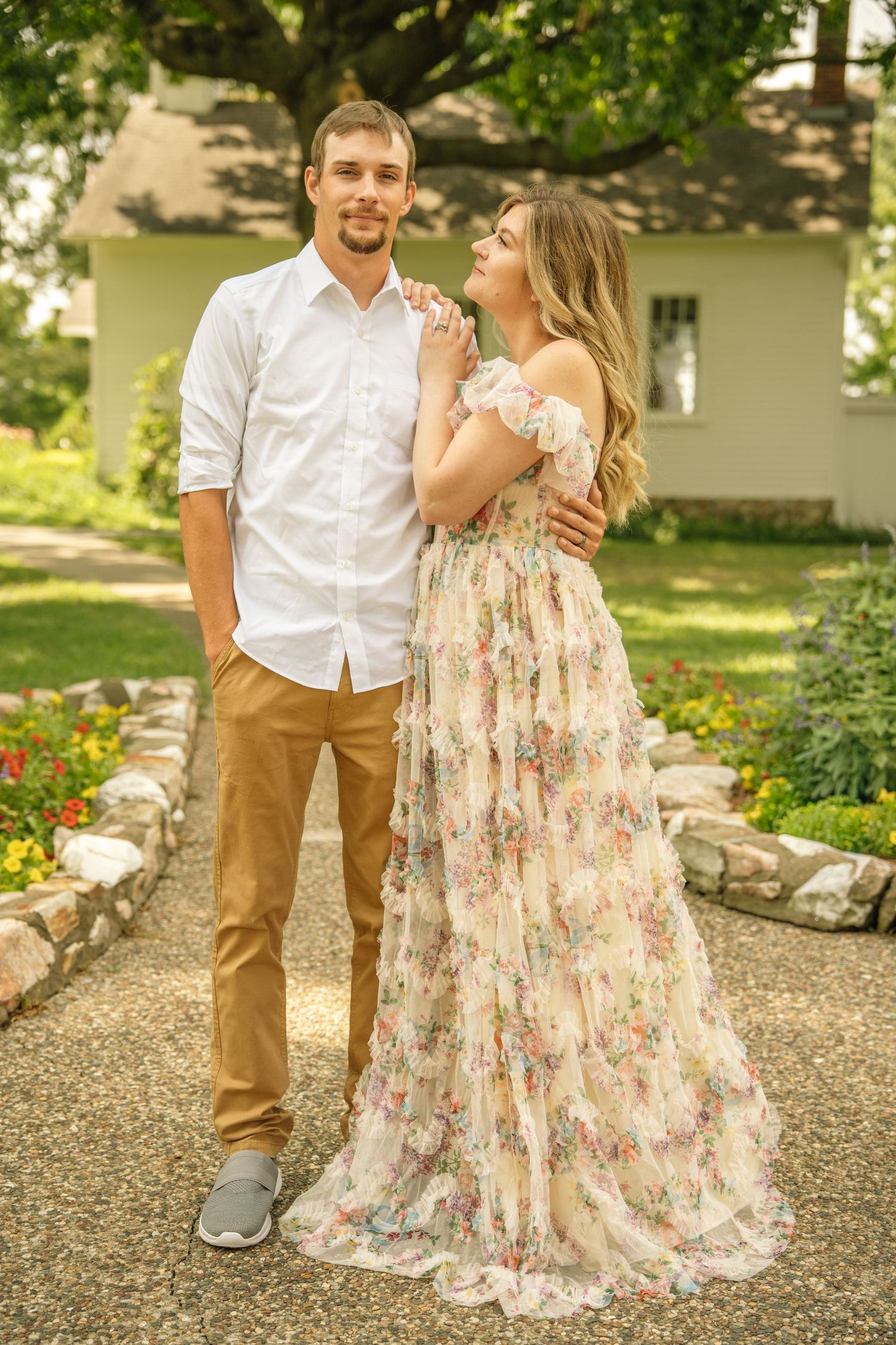 A young couple poses in a garden, the woman in a floral off-shoulder gown gazing at her partner near a white house.