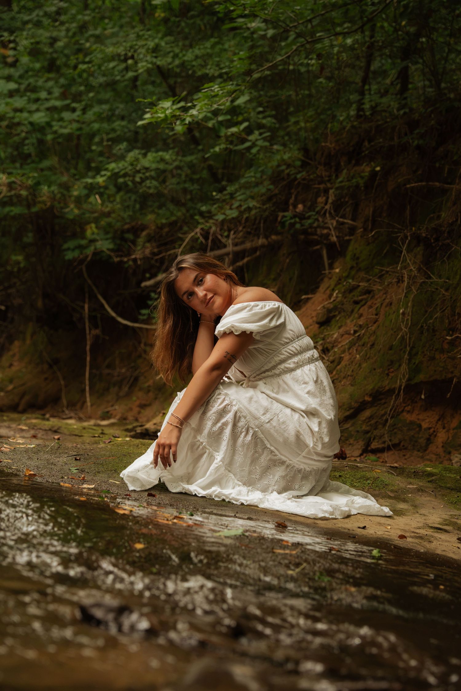 Woman in white dress crouches by rocky forest stream, touching water with serene expression.