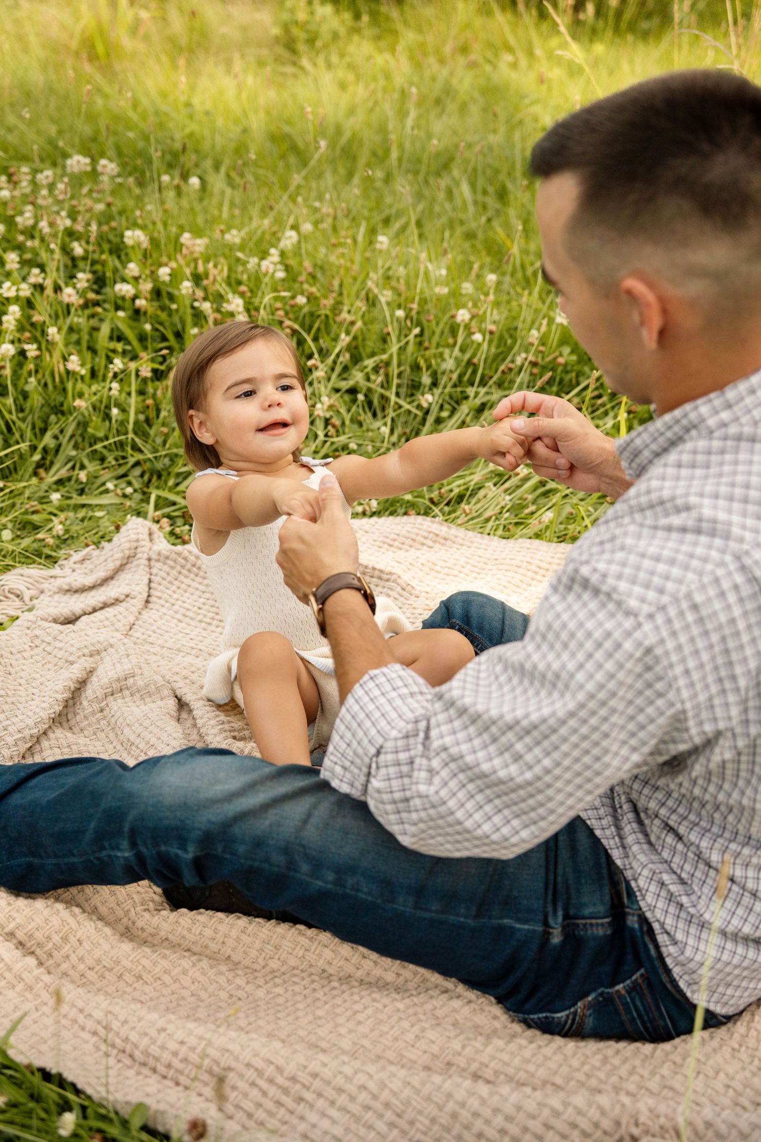 Father sitting on blanket in meadow playing with smiling toddler daughter outdoors.