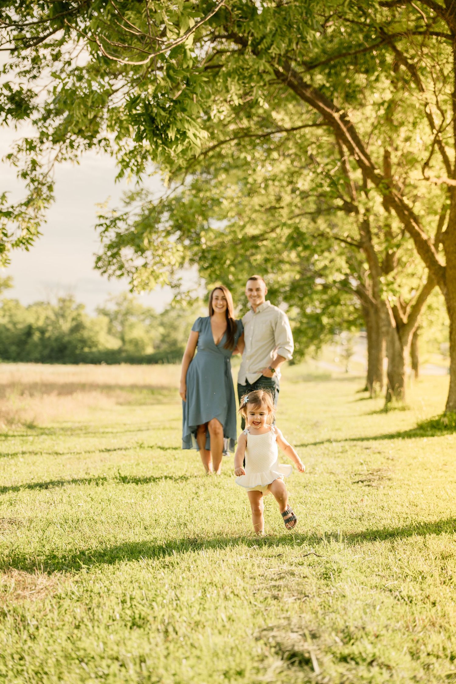 Happy family with toddler girl running in sunny green park, parents smiling in background among trees.