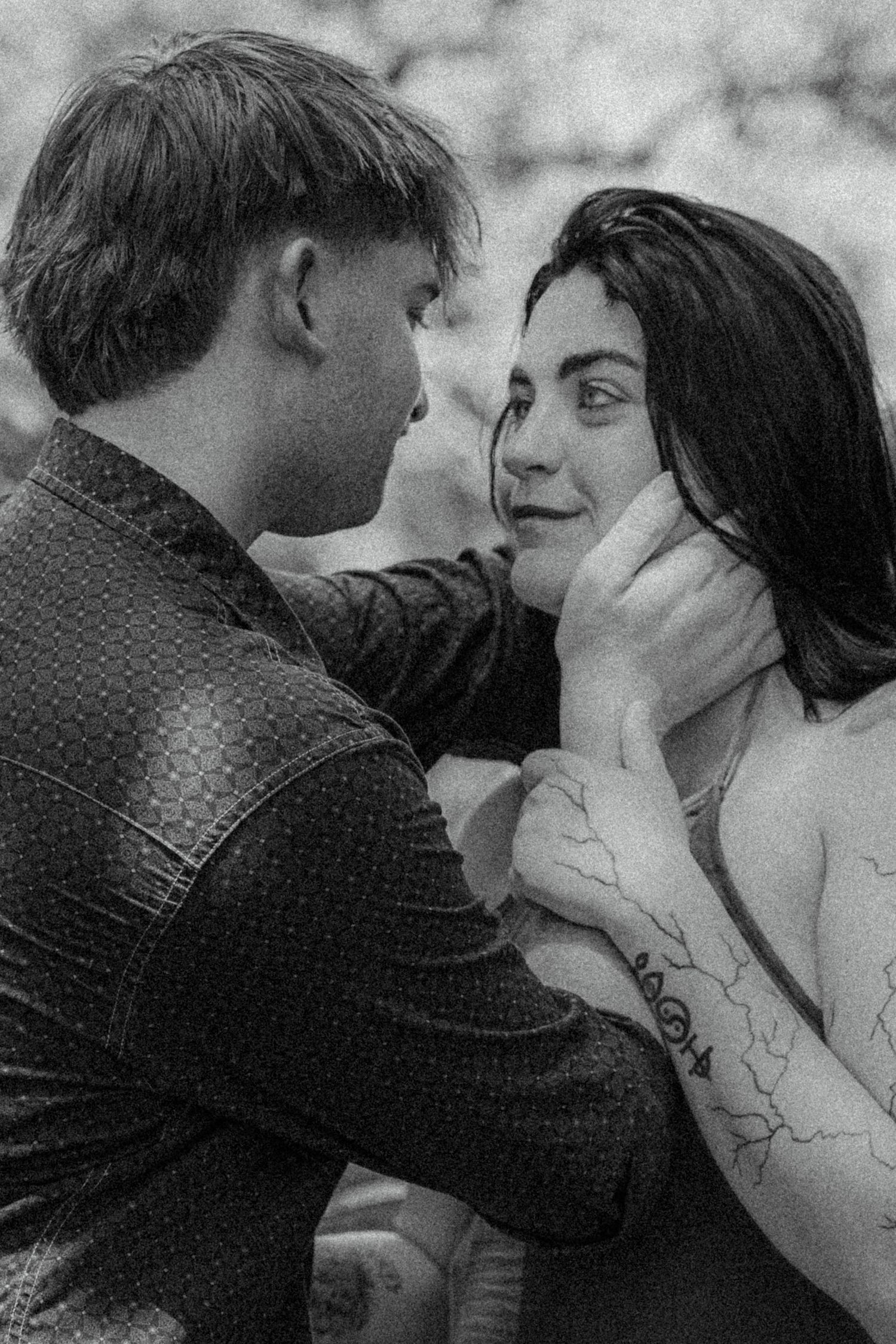 Black and white photo of a couple sharing an intimate moment, man gently holding womans face, tattoo visible on her arm.