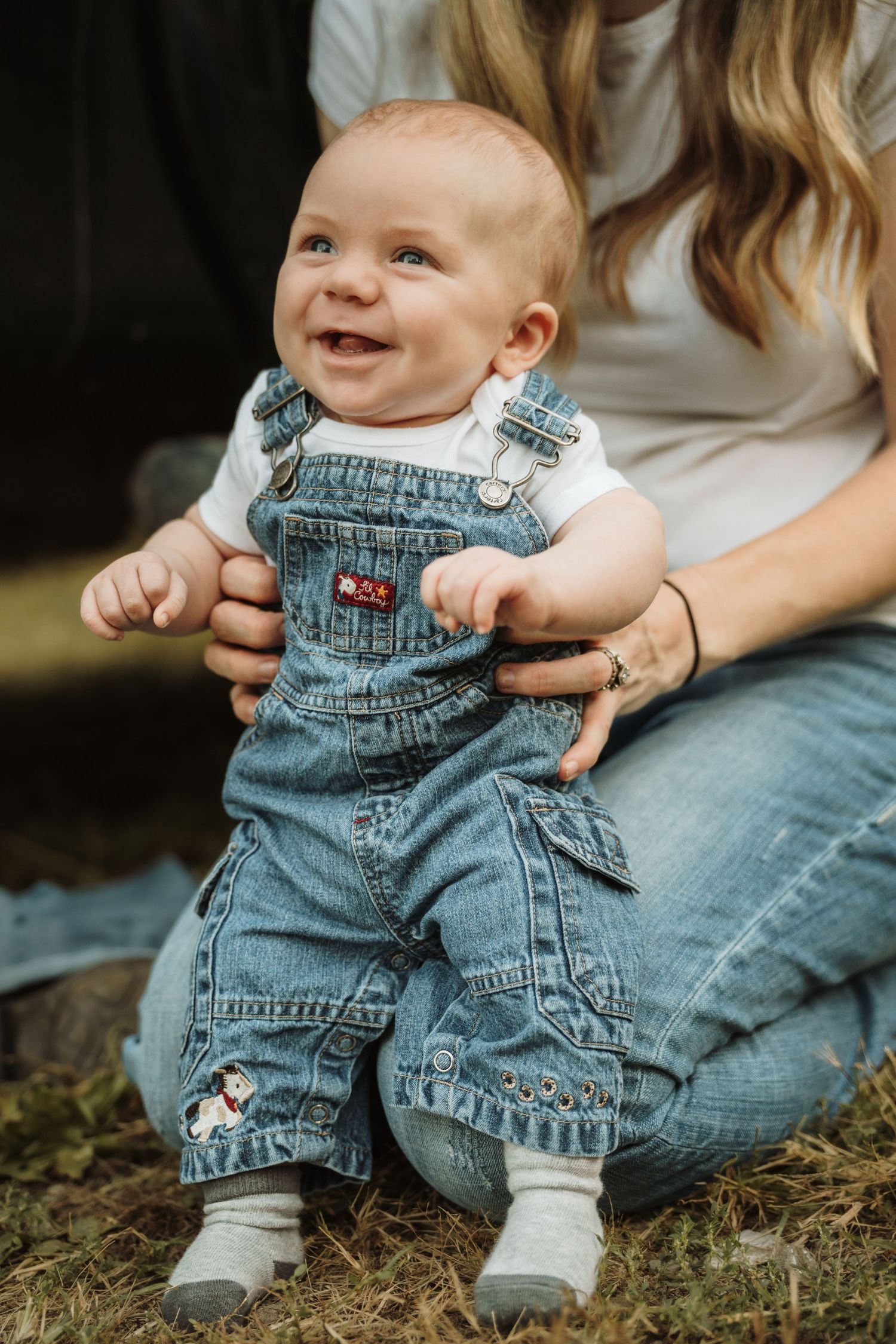 Smiling baby in denim overalls and white onesie sitting on mother's lap outdoors on grass.