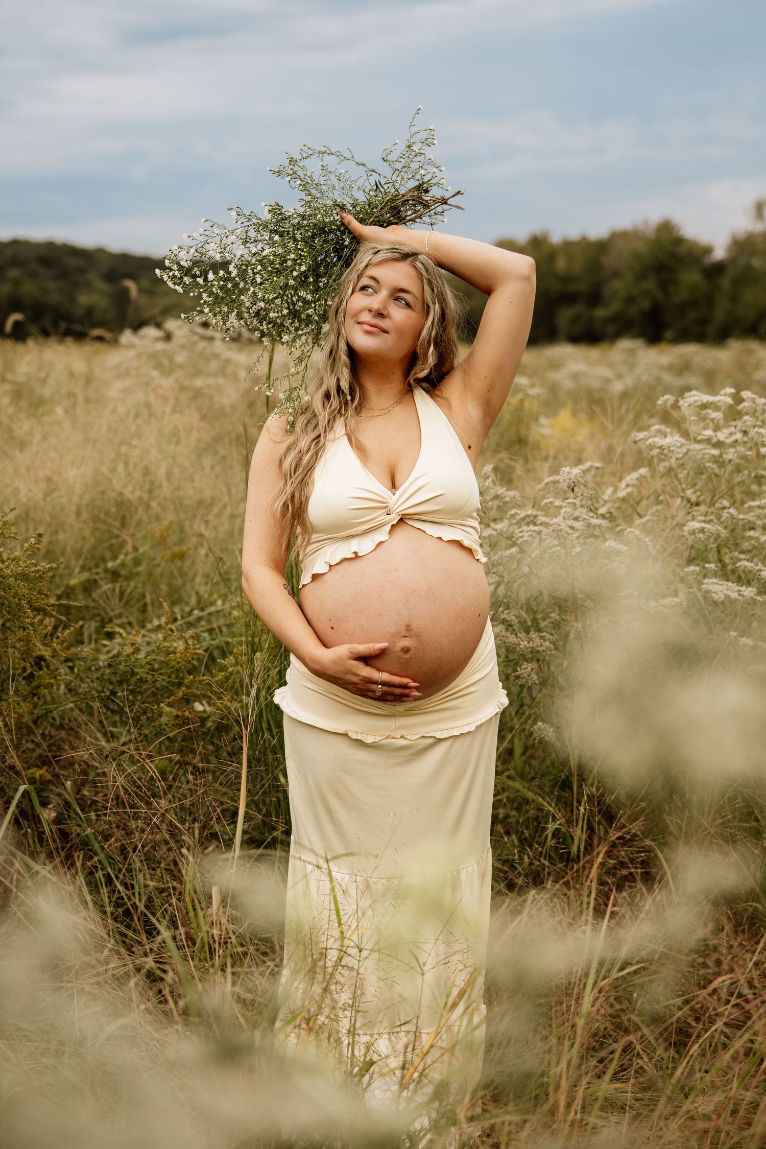 Pregnant woman in cream outfit holds wildflowers overhead while standing in a meadow field.