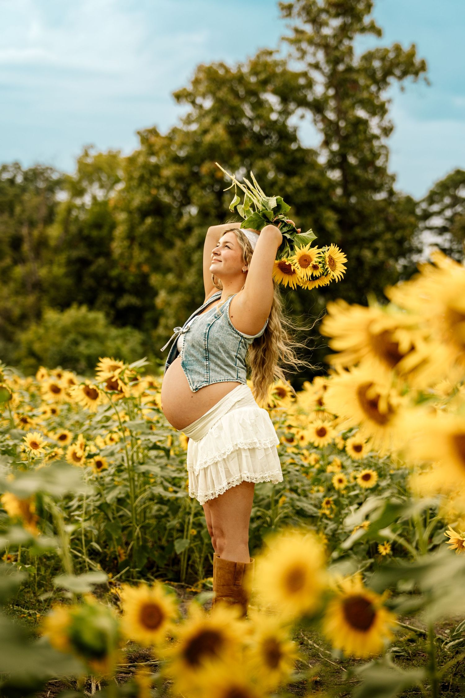 Pregnant woman in denim top and white skirt holding sunflowers in a vibrant sunflower field on a sunny day.