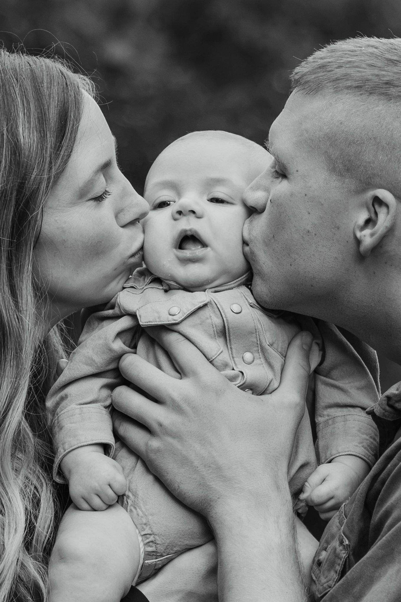 Black and white photo of parents kissing their newborn baby on the cheeks while holding them outdoors.