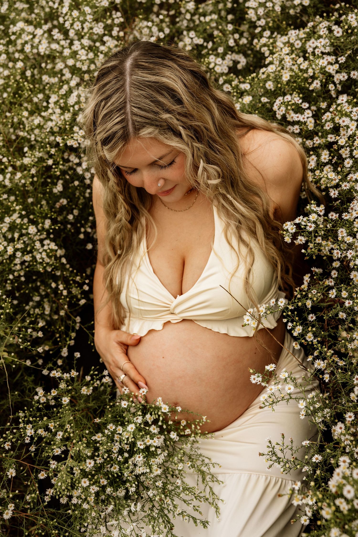 Pregnant woman in cream halter top cradles her belly surrounded by delicate white wildflowers.