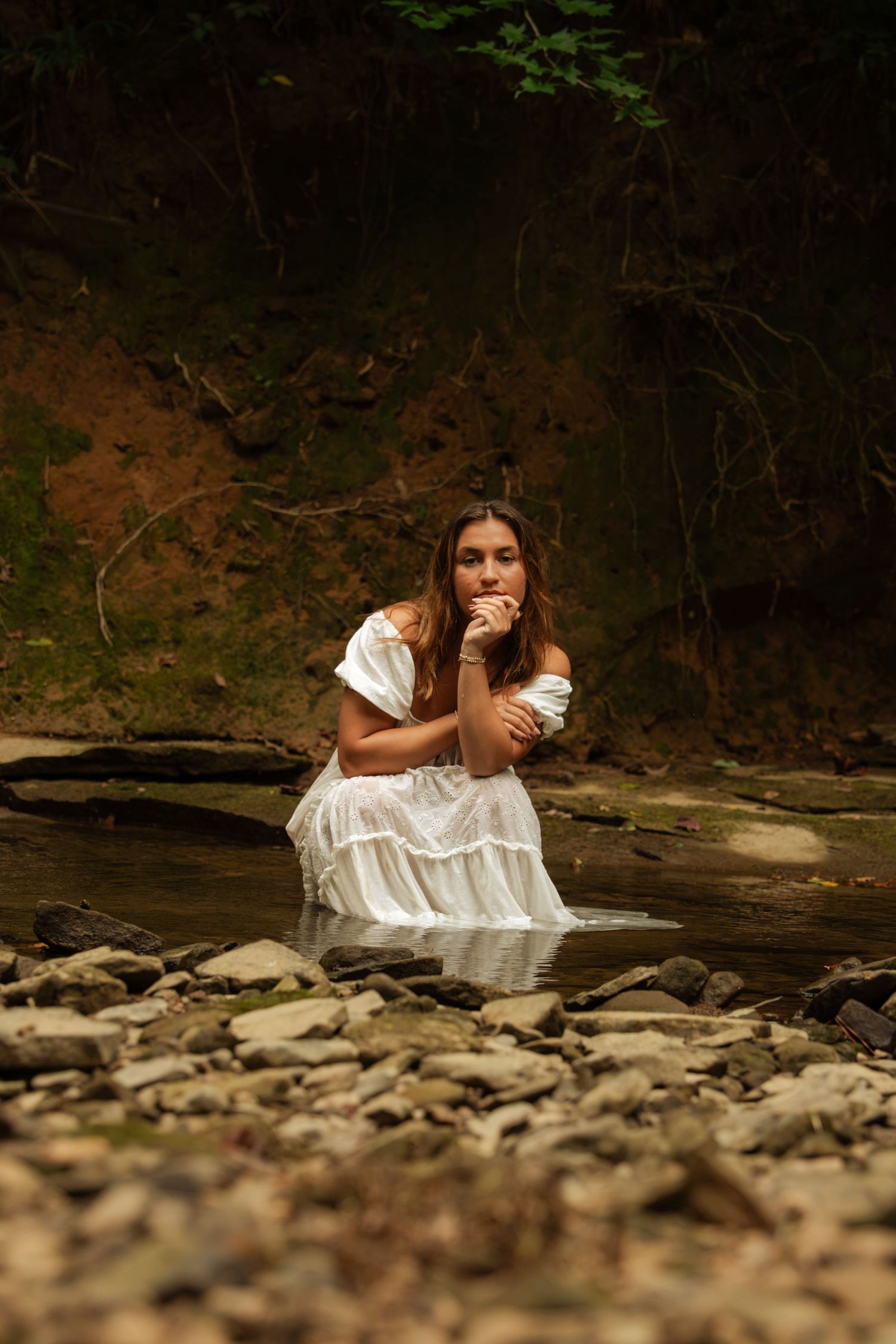 Woman in white dress sitting in rocky stream surrounded by dark forest backdrop.