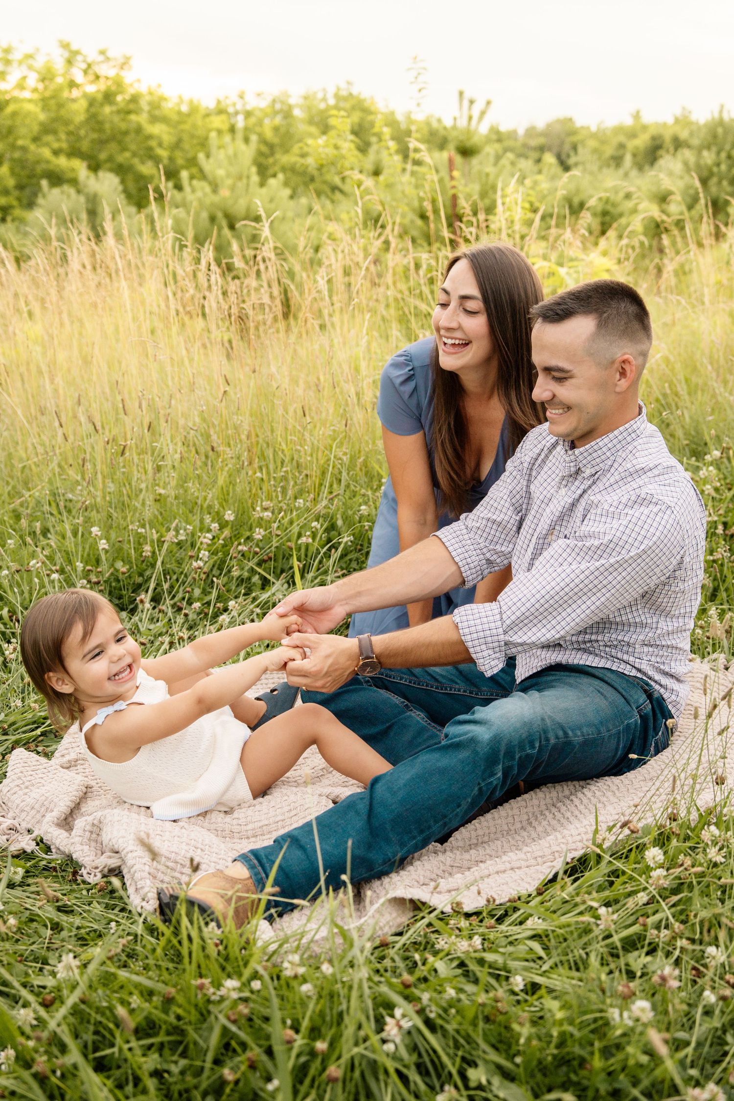 Happy young family sitting on a blanket in a sunny green field, playing with their smiling toddler daughter.