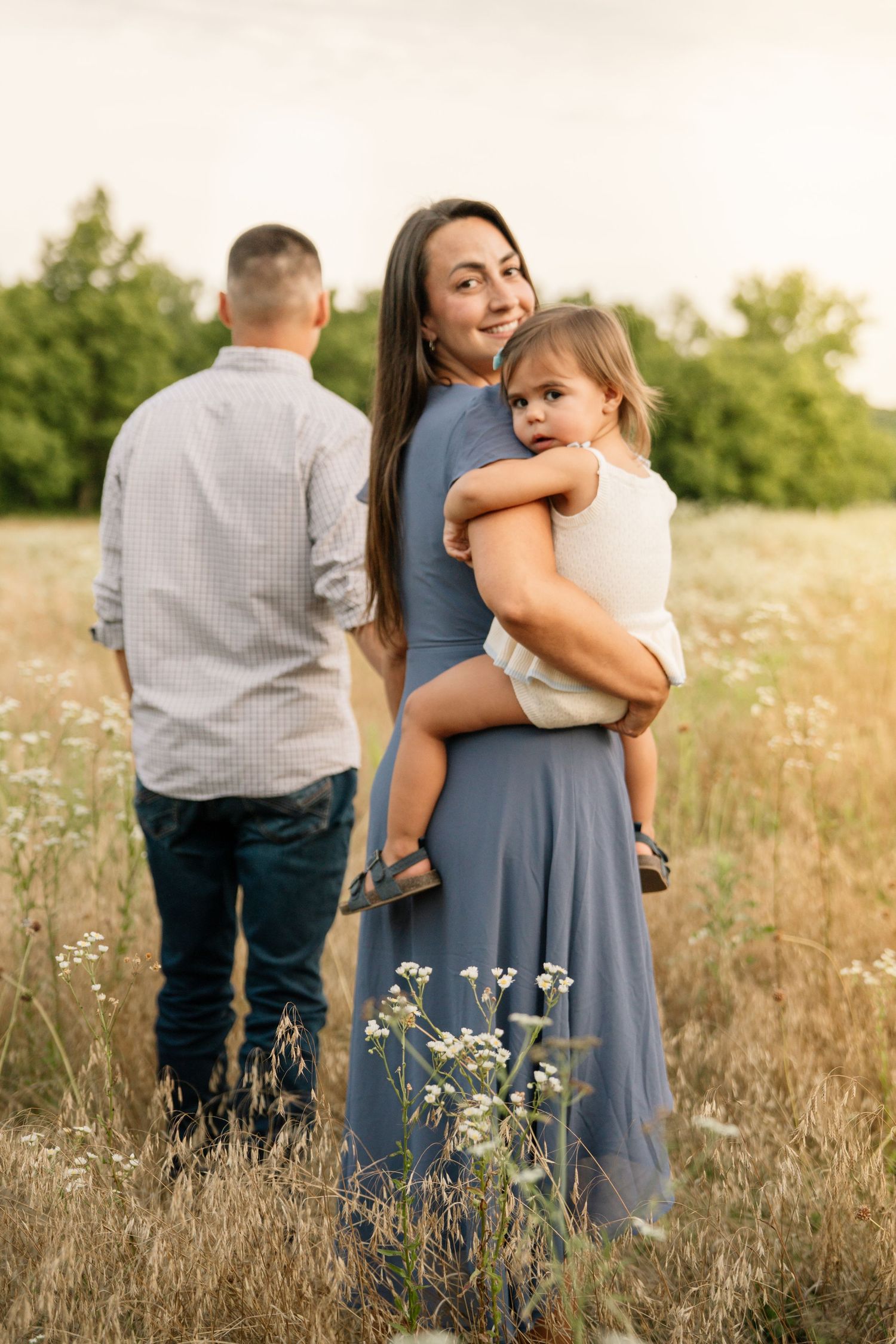 Smiling mother in blue dress holds young child as father stands in golden meadow.