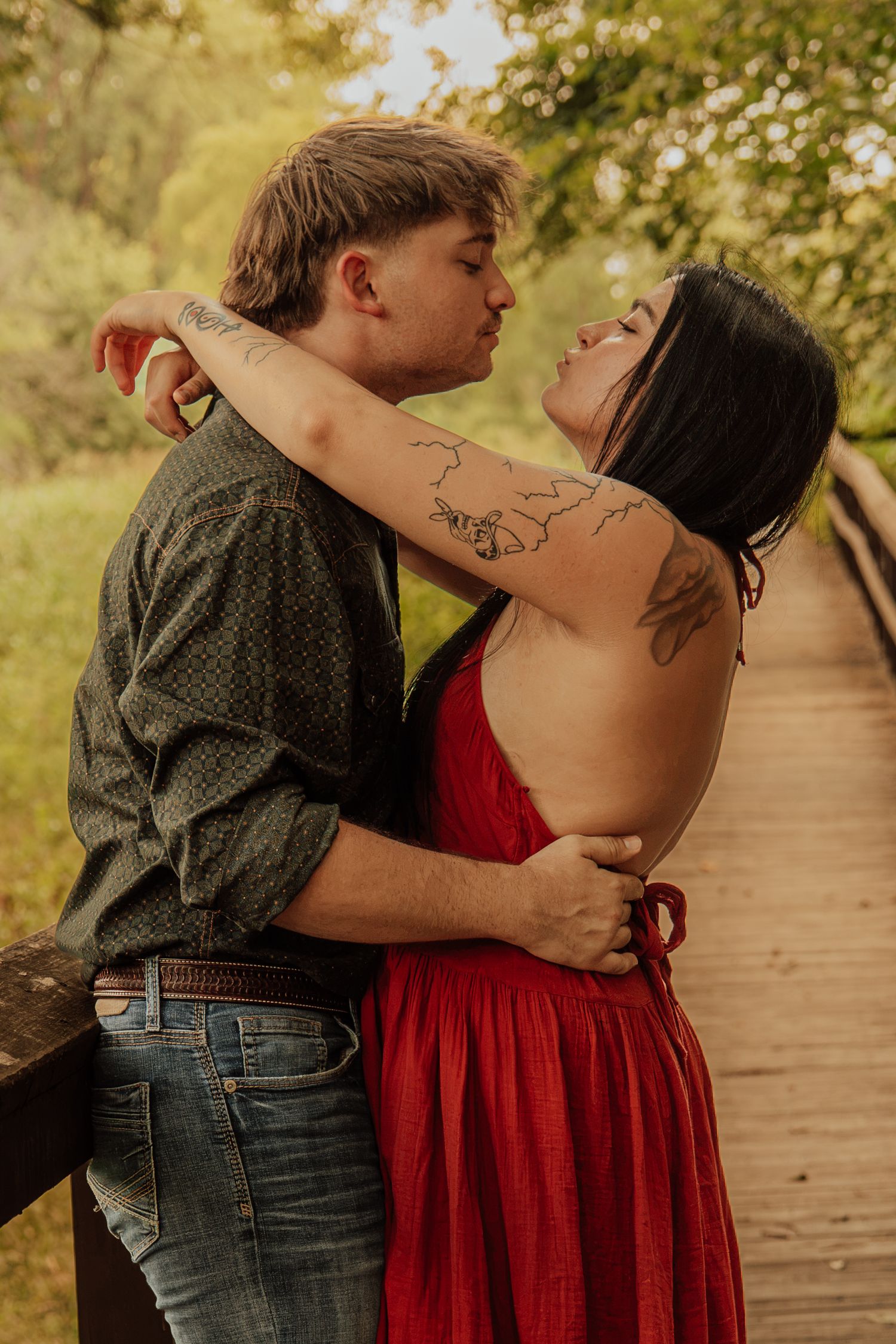 A couple embraces on a wooden bridge, the woman in a red dress with tattoos leaning back as her partner kisses her cheek.