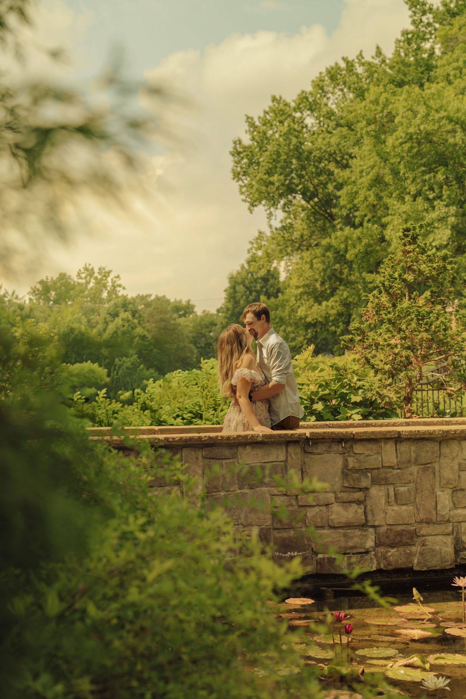 Romantic couple sharing a kiss on a stone wall bridge surrounded by lush greenery and lily pads.