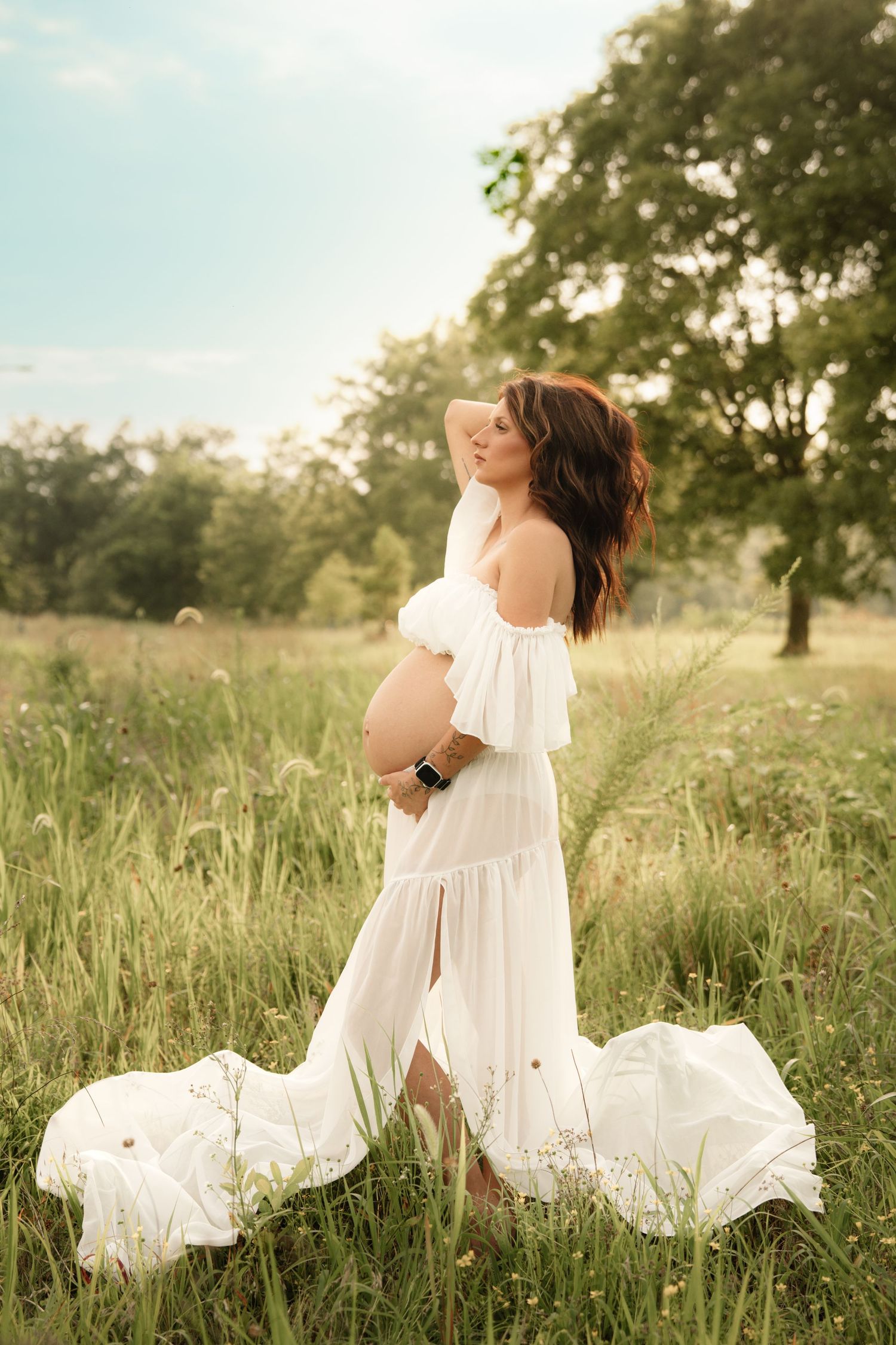 Pregnant woman in flowing white maternity dress stands in a sunlit meadow with fabric trailing on grass.