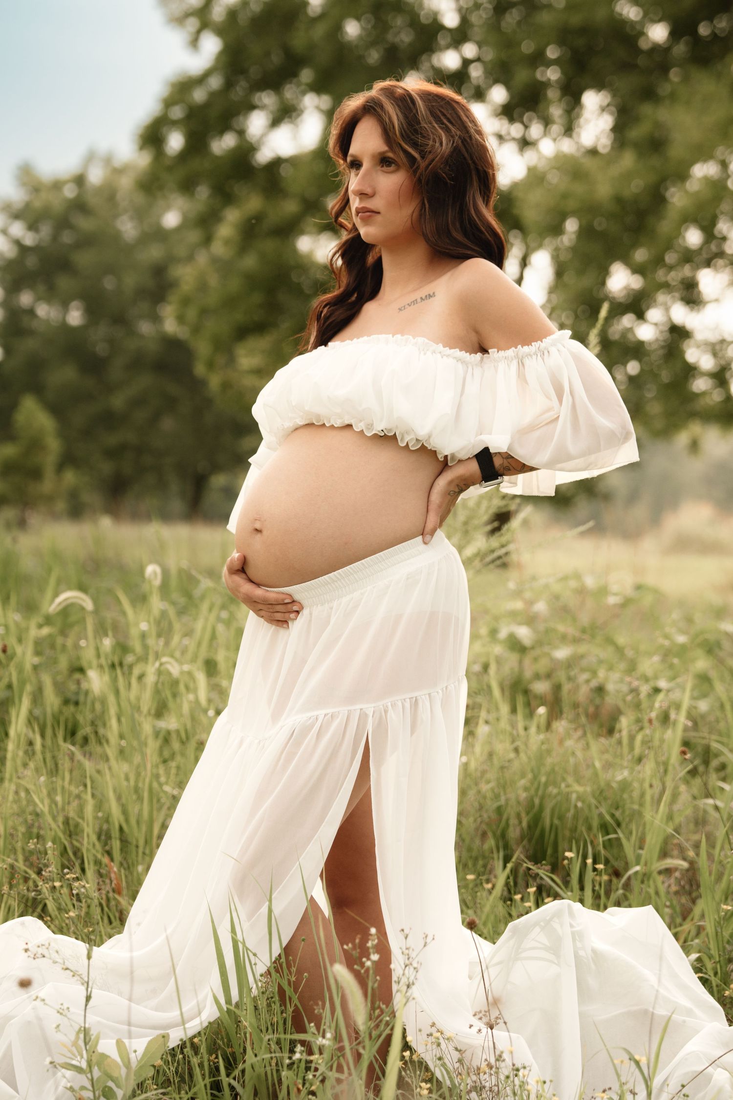 Pregnant woman in white off-shoulder dress stands in wildflower field holding baby bump confidently.