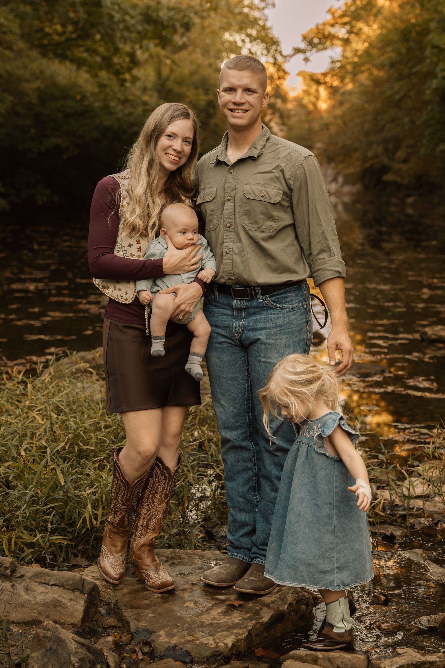Family of four poses on rocks by a creek in golden autumn light, mom holding baby and toddler girl nearby.