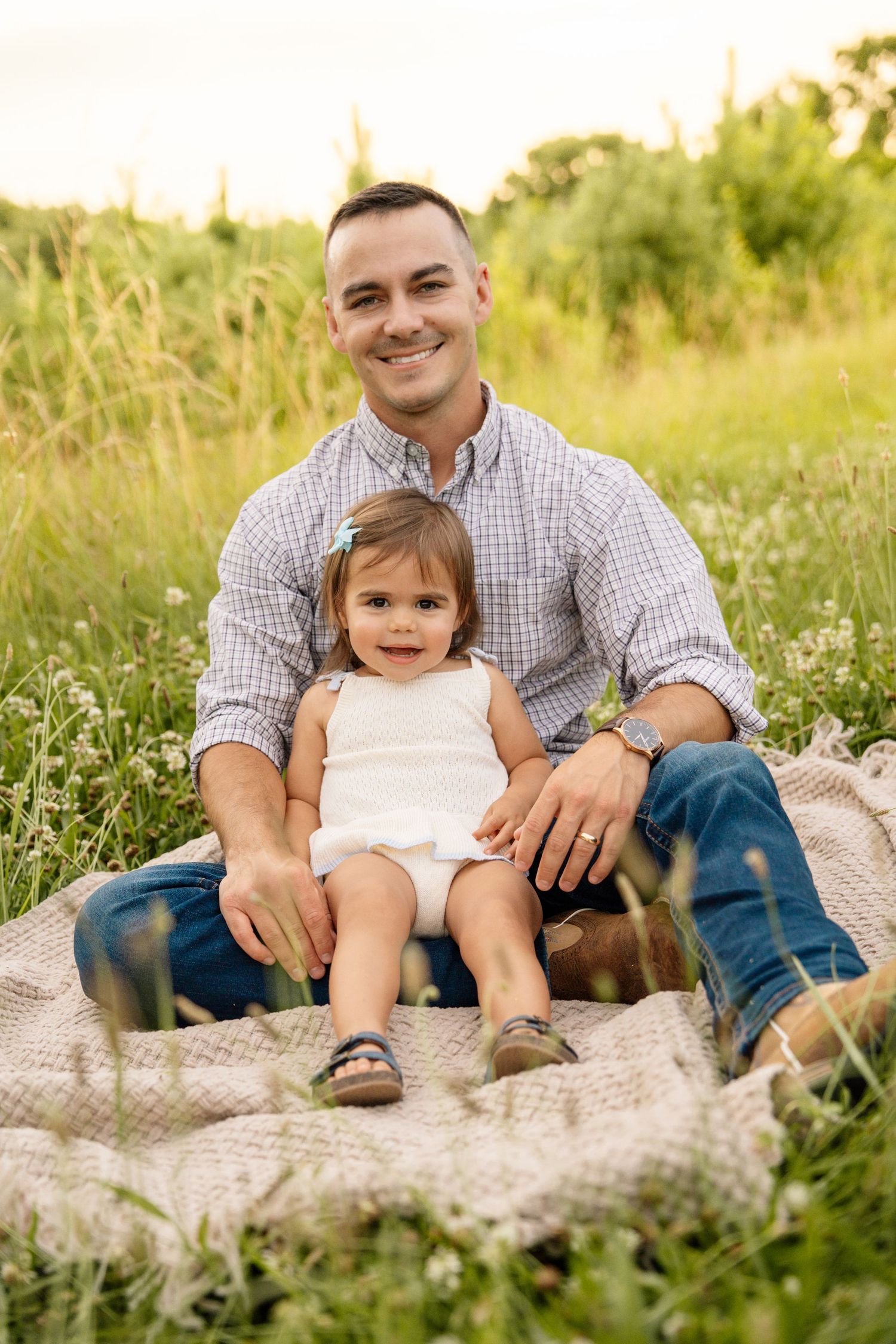 Smiling father sitting on a blanket in a green meadow with his toddler daughter between his legs.