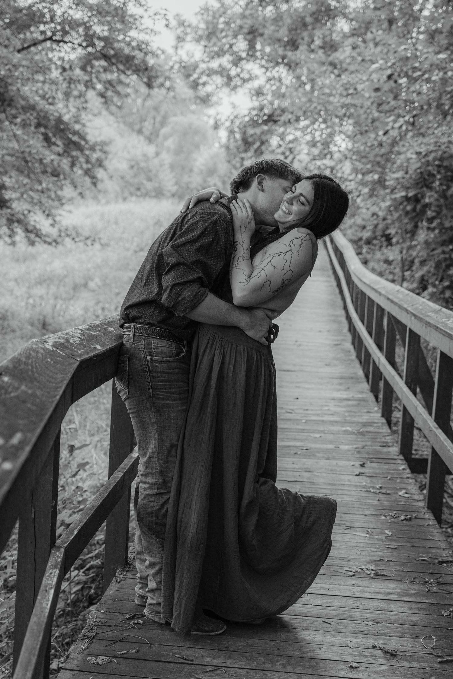 Black and white photo of a romantic couple kissing on a wooden bridge surrounded by lush trees.
