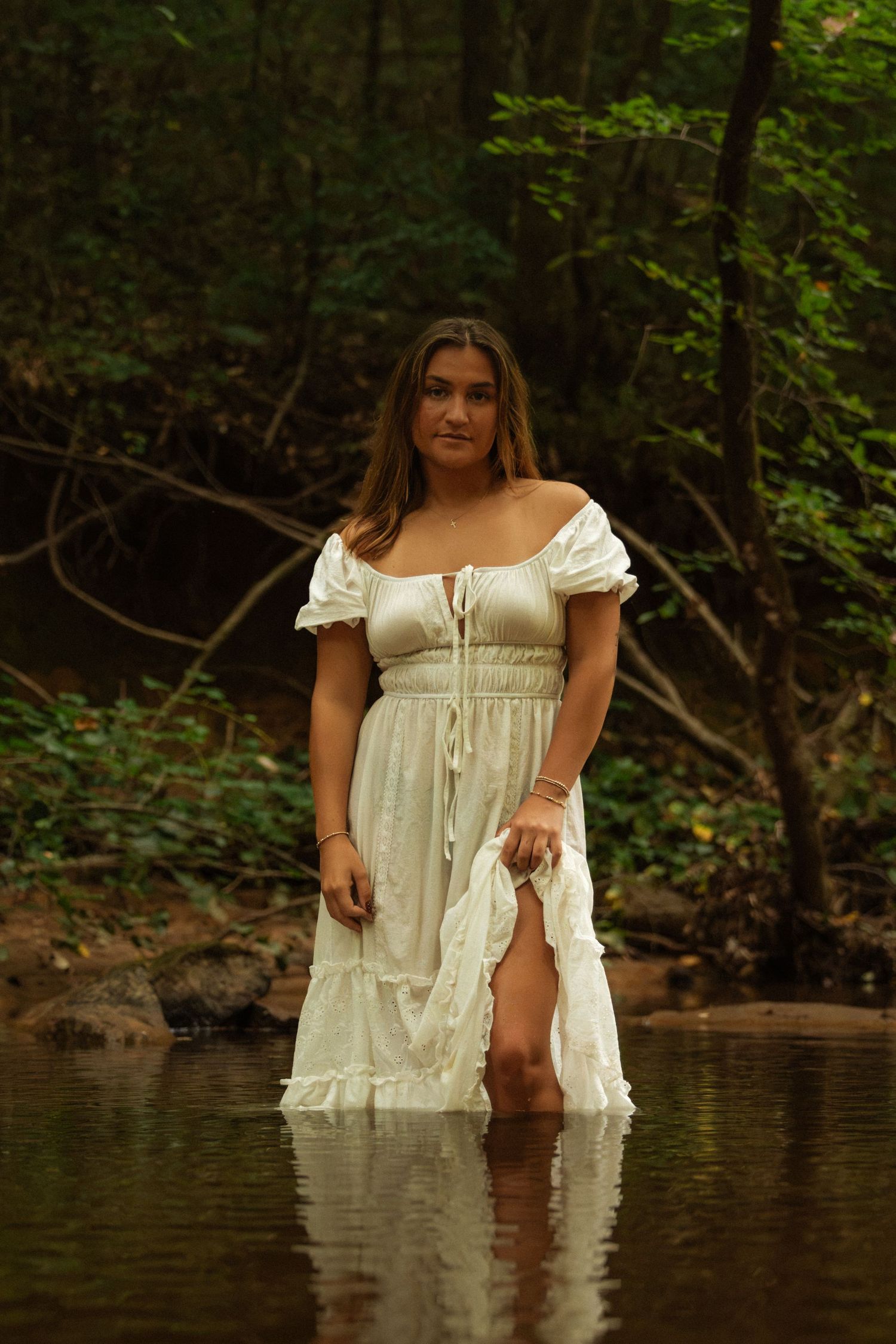 Woman in white off-shoulder dress stands in shallow forest creek, surrounded by lush trees.