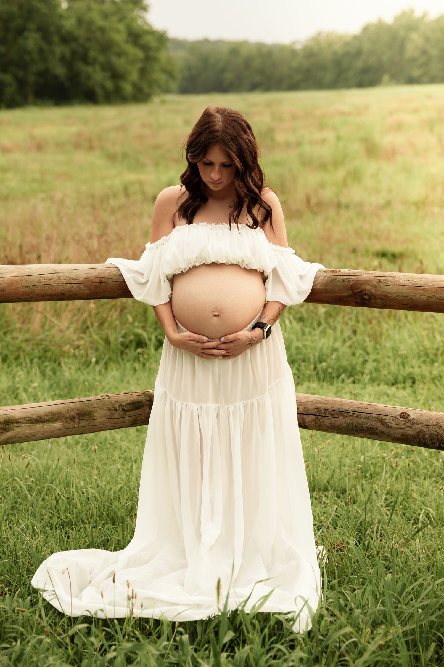 Pregnant woman in flowing white dress stands by rustic wooden fence in green meadow holding bump.