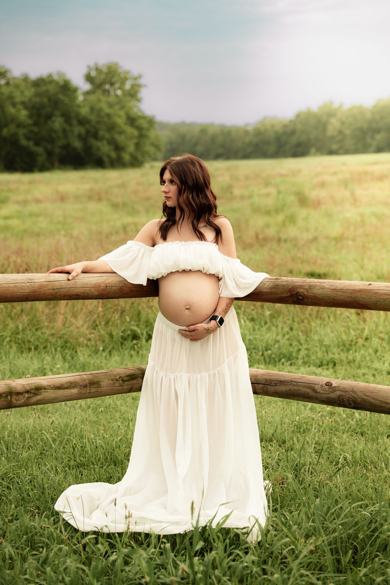 Pregnant woman in white off-shoulder dress cradles her baby bump leaning against a rustic wooden fence.