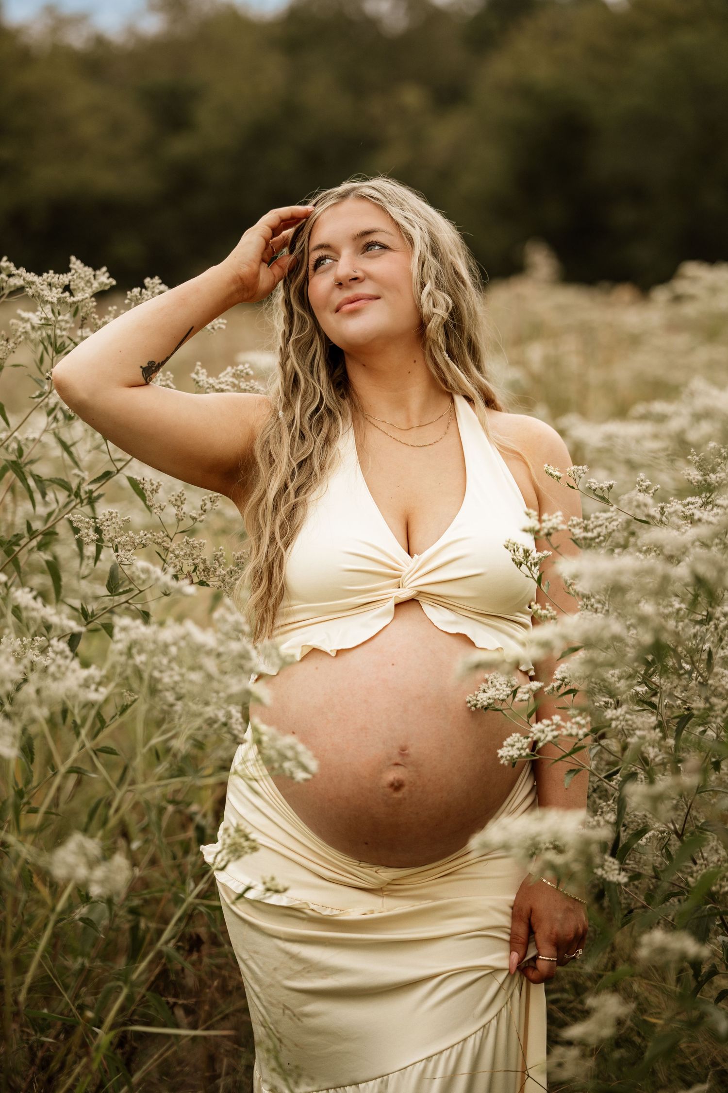 Pregnant woman in yellow crop top and skirt standing in a field of white wildflowers, smiling outdoors.