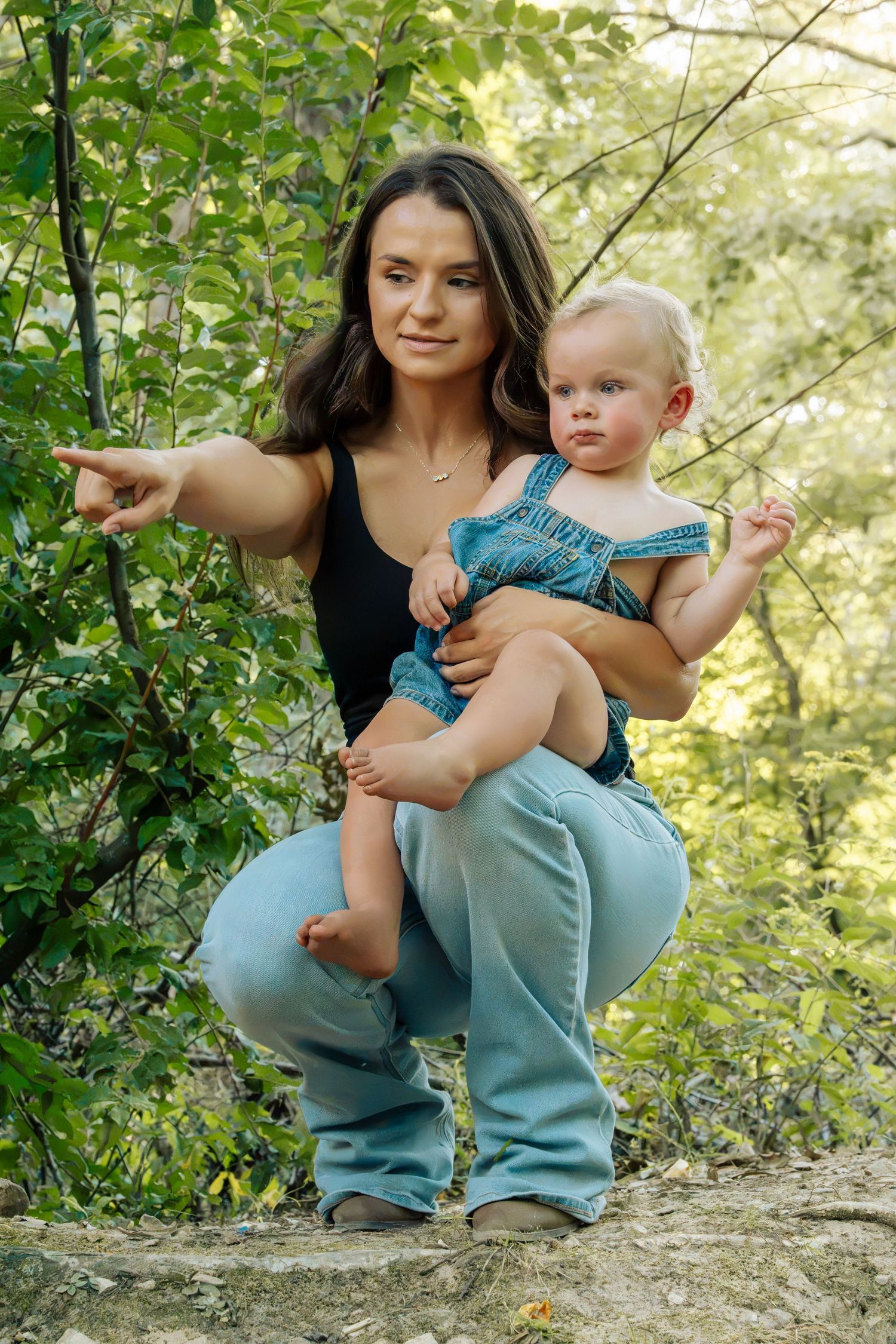 Mother crouching with baby in denim overalls, pointing in lush green forest.