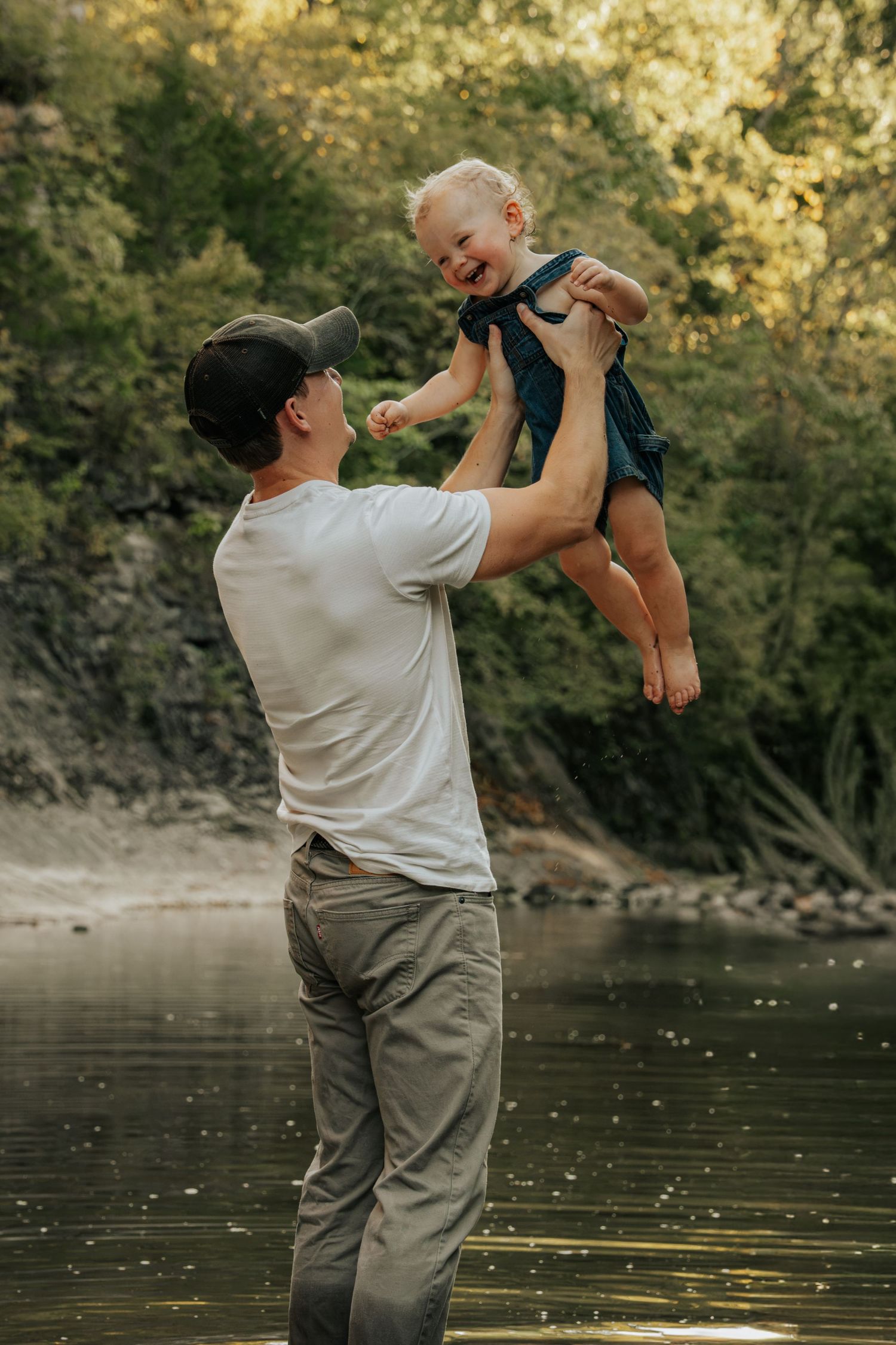 Joyful father lifting laughing toddler in denim overalls high in the air by a scenic river.