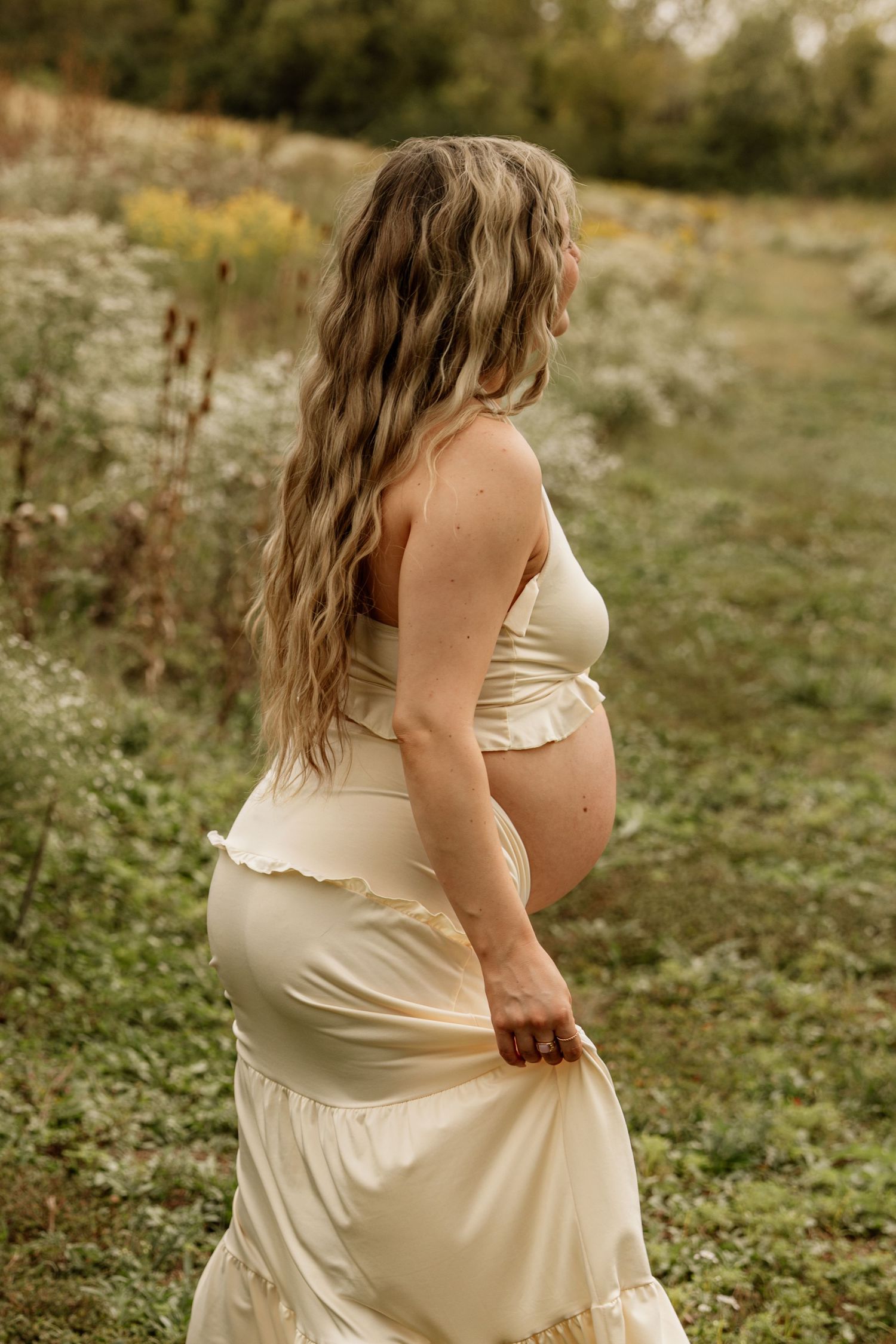 Pregnant woman in cream two-piece maternity dress standing in a lush green field with wildflowers.