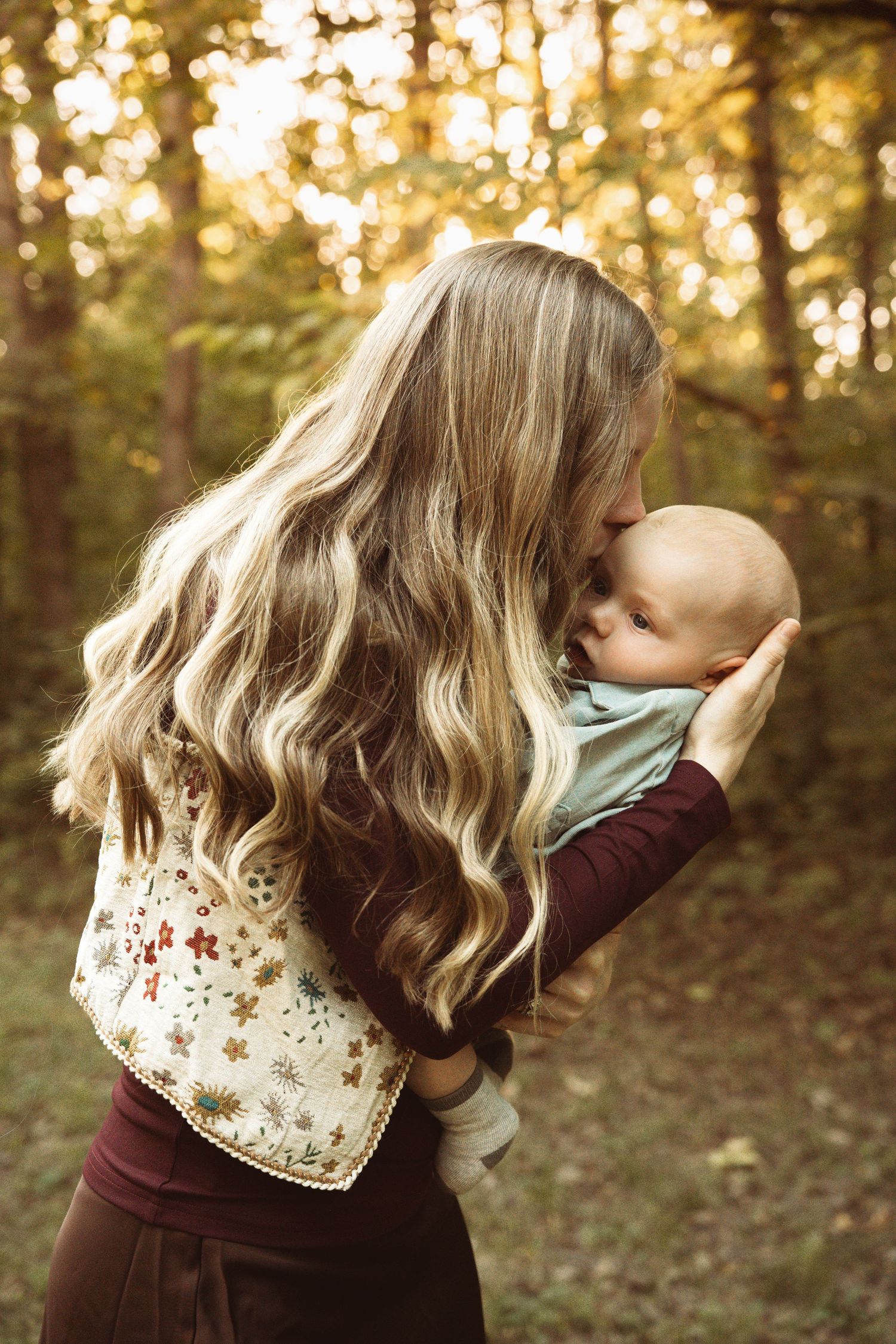 Mother with long wavy blonde hair kissing her newborn baby outdoors in a sunlit forest setting.