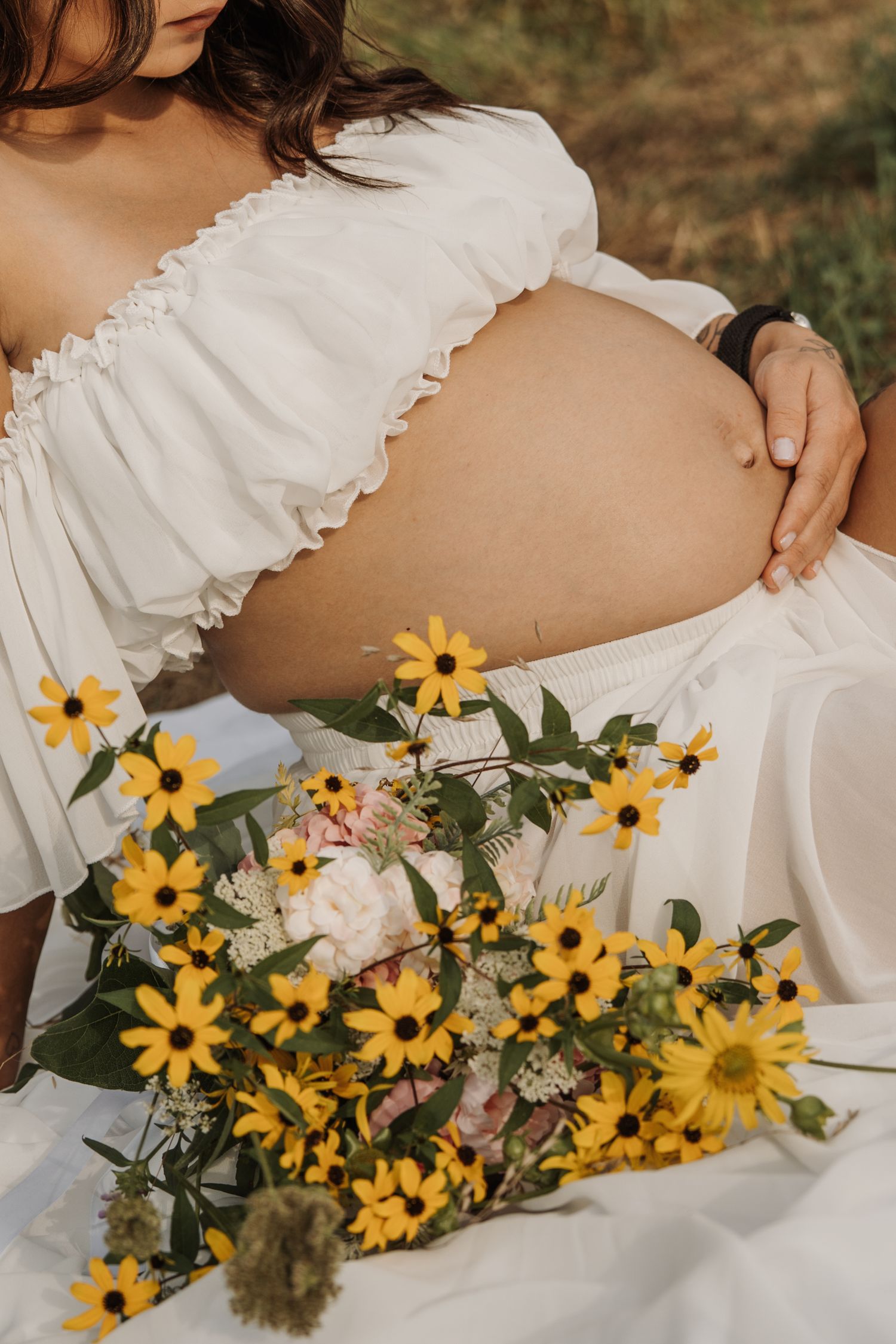 Pregnant woman in white off-shoulder top holds baby bump surrounded by yellow wildflowers outdoors.