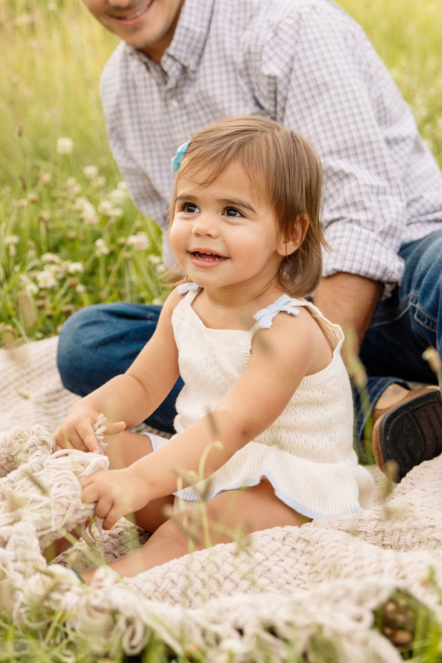 Happy toddler girl in white romper sitting on a blanket in a wildflower field smiling brightly.