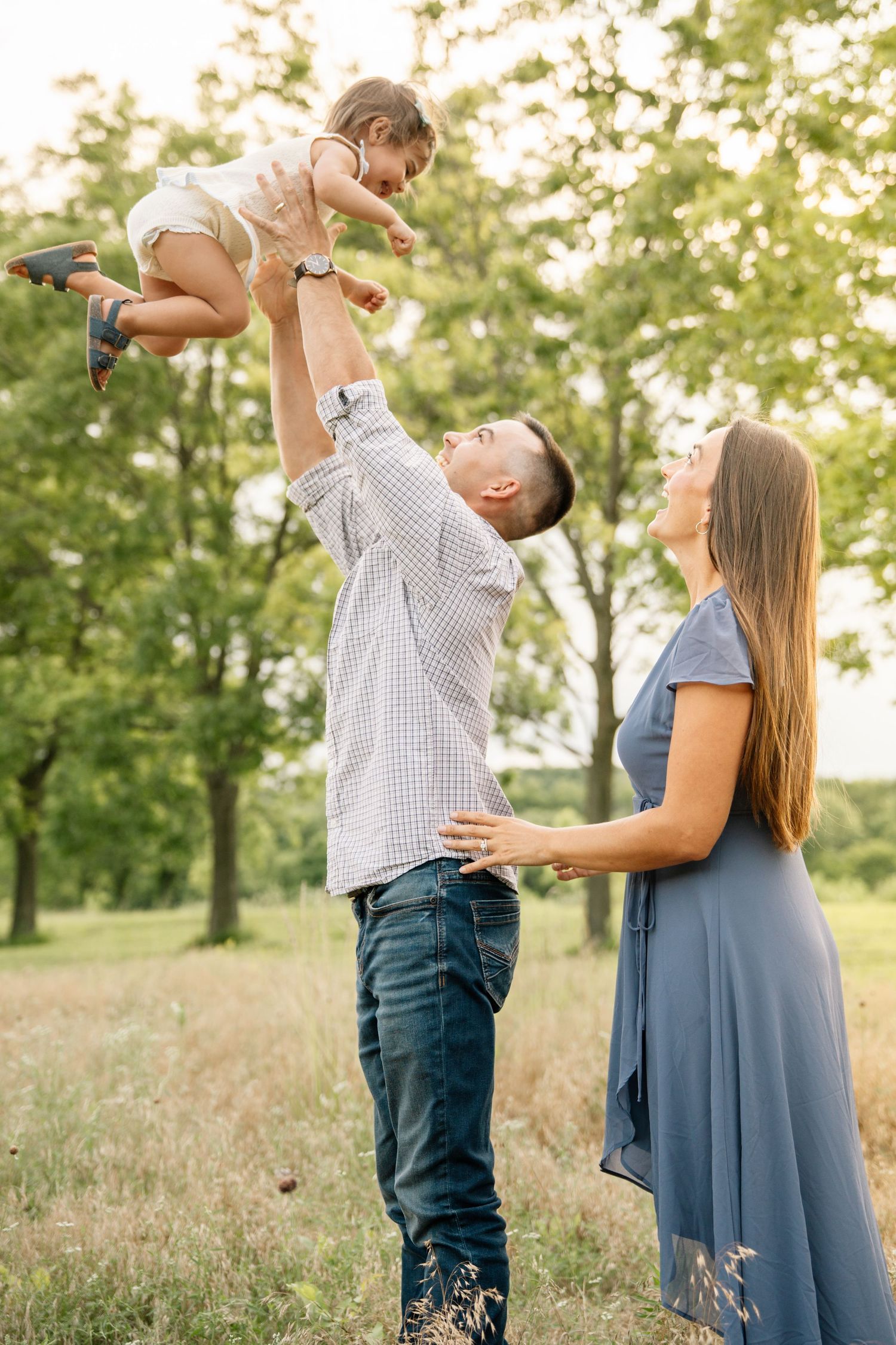 Happy family outdoors, father lifting baby in air while mother watches, surrounded by green trees.