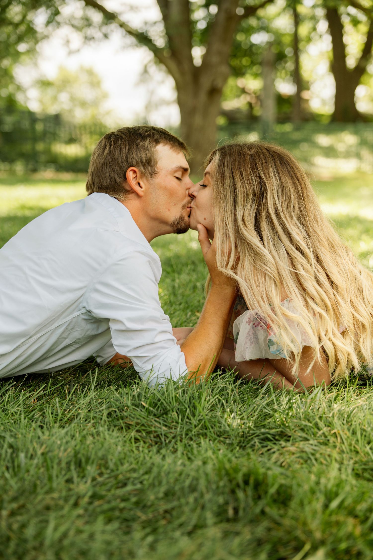 Couple sharing a tender kiss while lying on green grass beneath a large tree in a sunlit park setting.