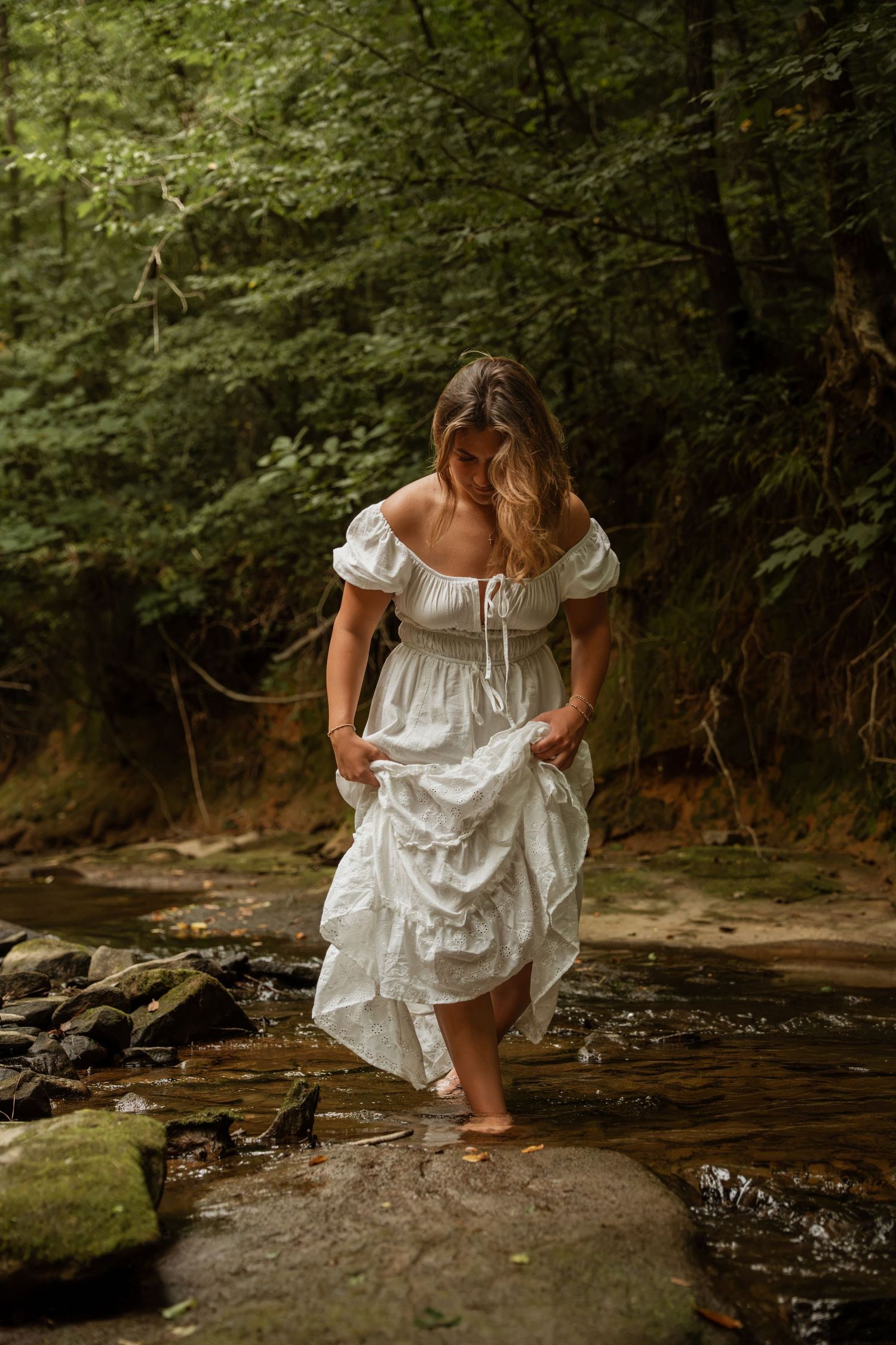 Woman in white off-shoulder dress lifting hem while walking barefoot on rocks by a forest stream.
