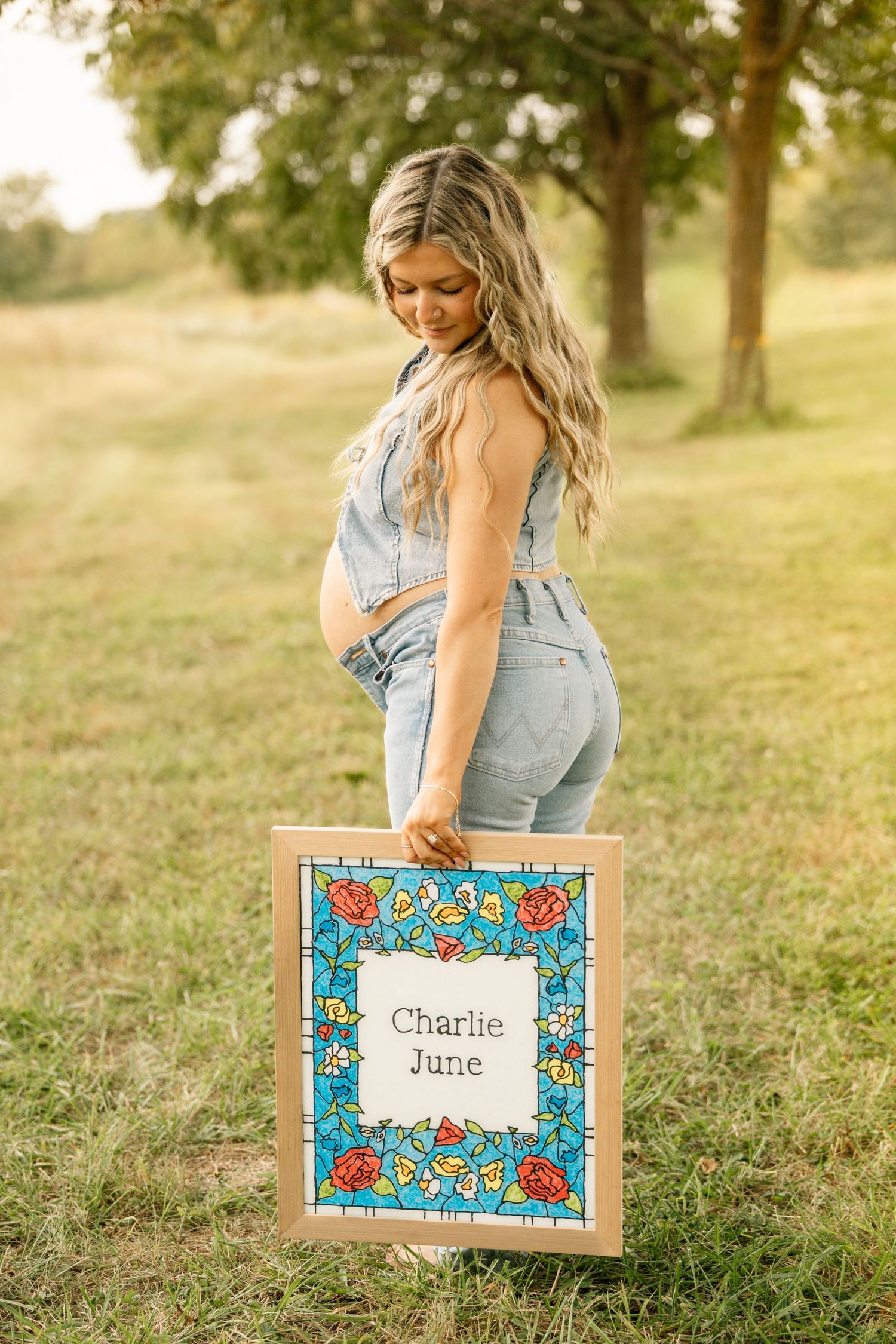 Pregnant woman in denim overalls holds a floral framed sign reading Charlie June in a grassy park.
