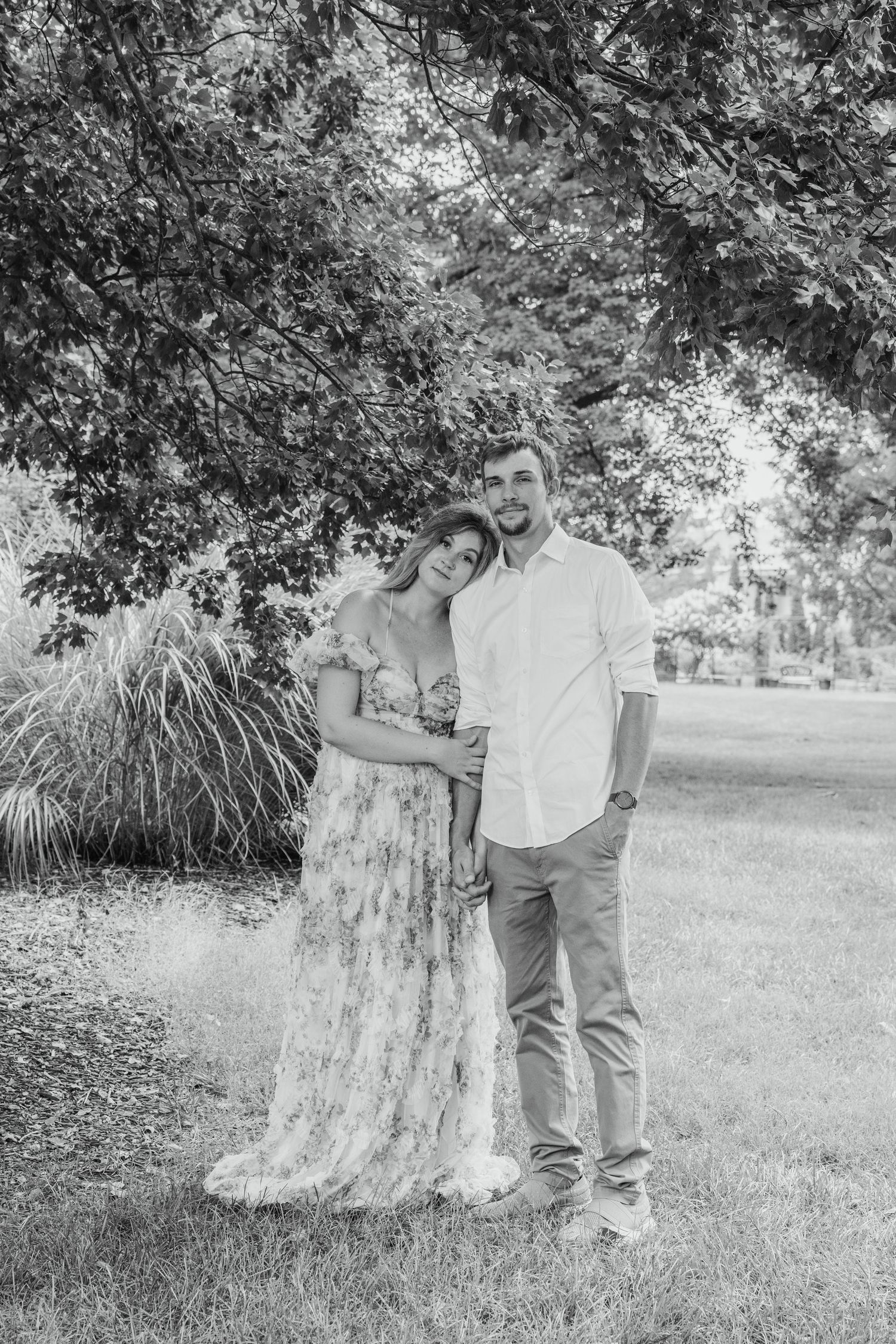 Black and white photo of a couple embracing outdoors under a large tree, woman in floral dress, man in white shirt.