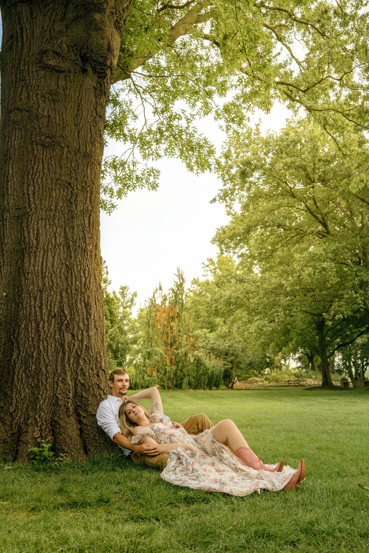 Couple relaxing under a large tree on green grass in a sunny park, woman in floral dress leaning on man.