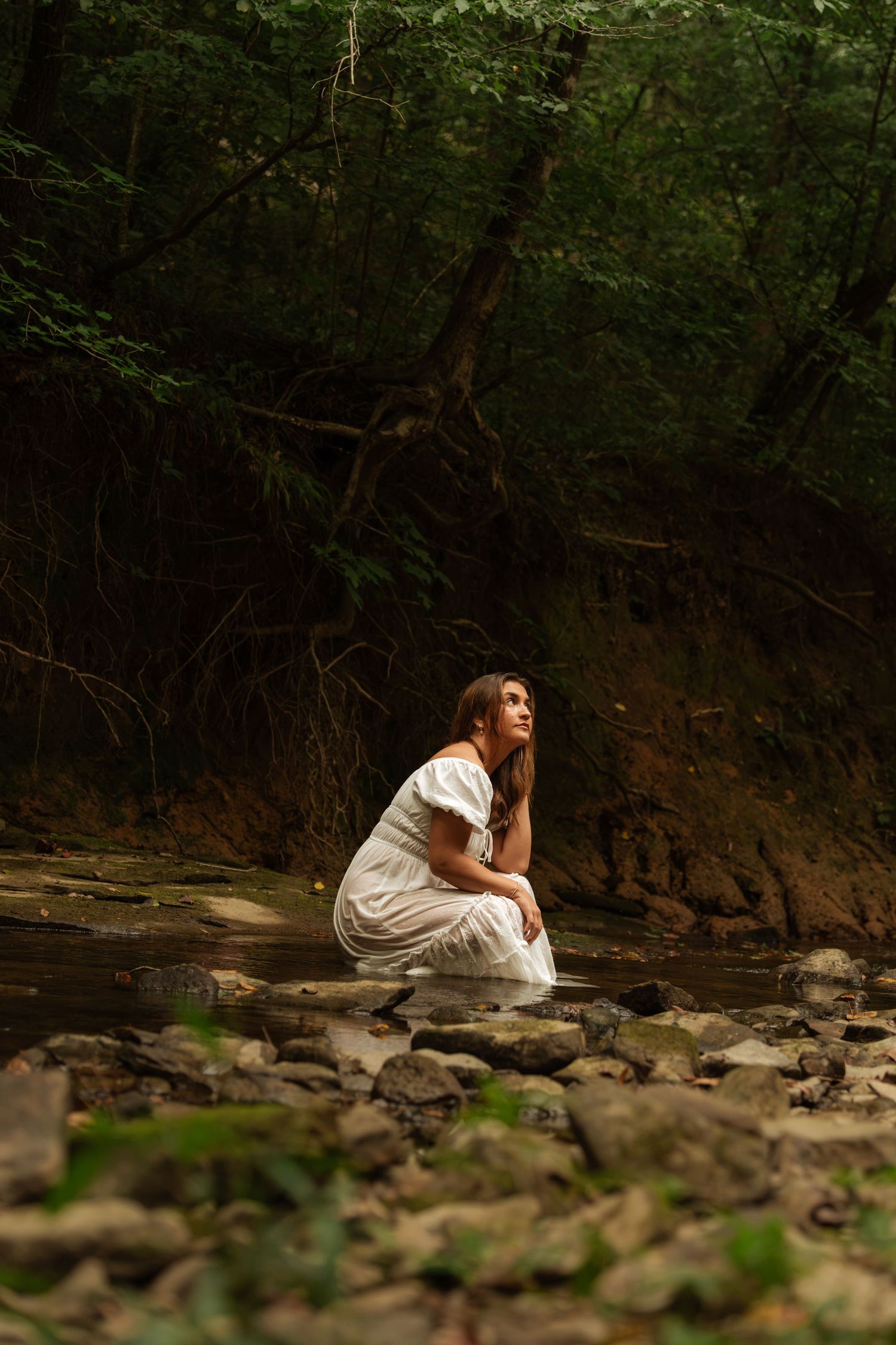Woman in white dress crouching on rocks by a forest stream, gazing upward in a serene woodland setting.