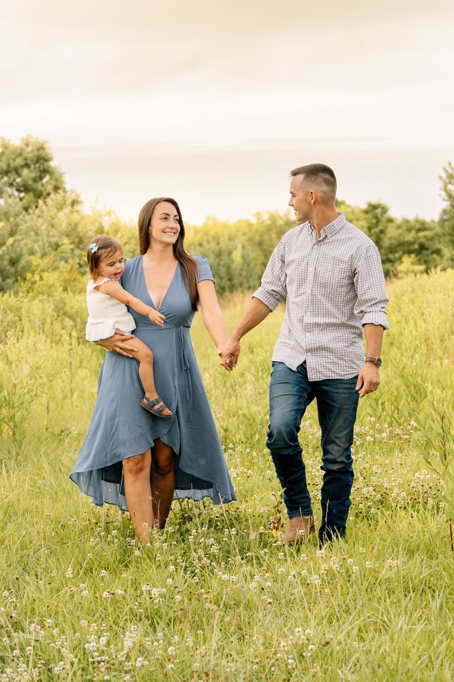 A happy family walks through a green meadow, mother in blue dress holding toddler, father in jeans holding hands.