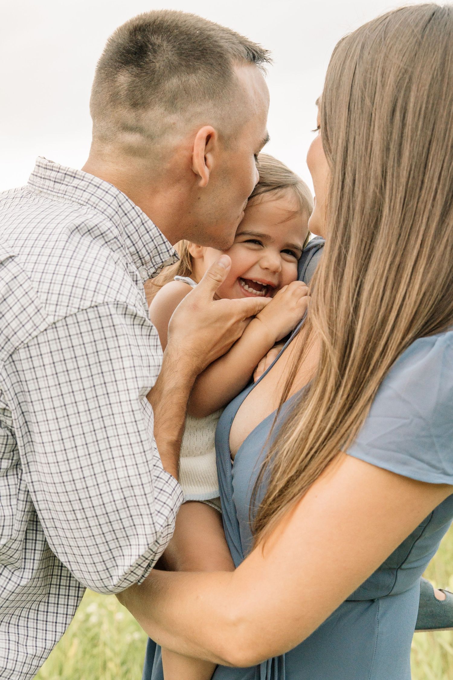 Happy family moment as parents kiss and cuddle their laughing toddler outdoors in a bright, natural setting.