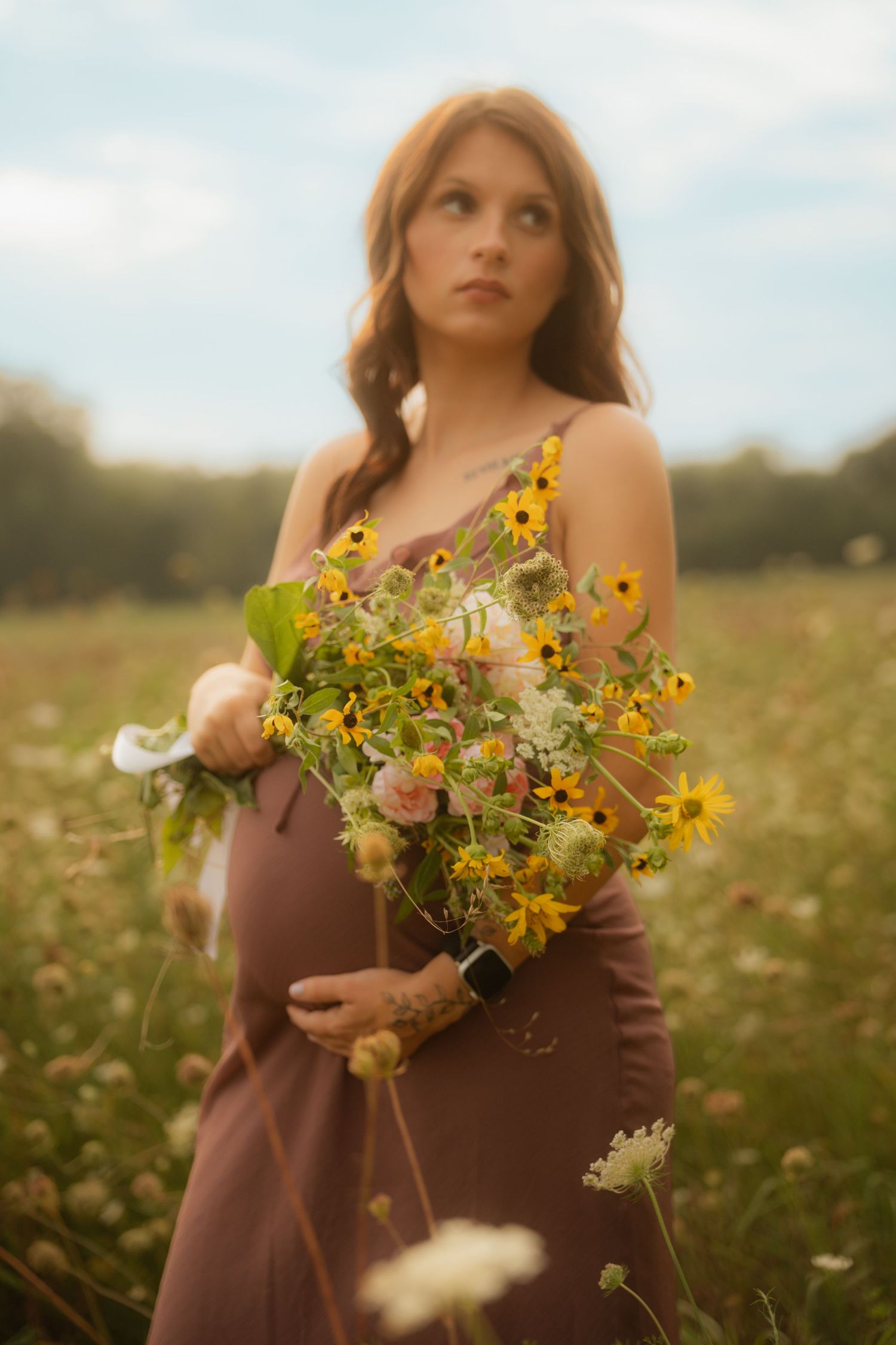 Pregnant woman in brown dress holding colorful wildflower bouquet in sunlit field, gazing upward thoughtfully.