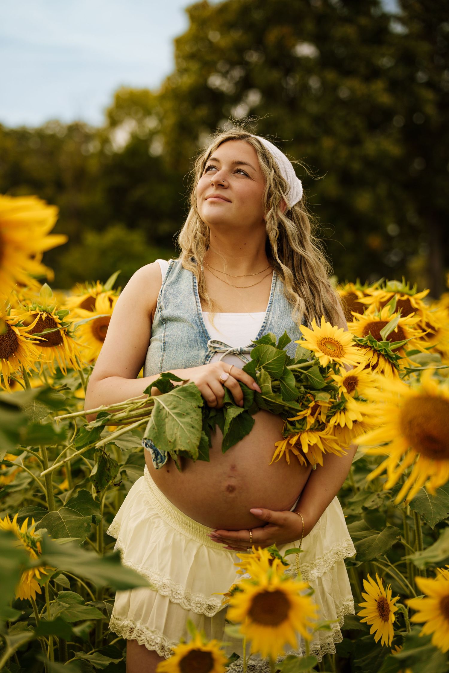 Pregnant woman holding sunflowers in a field, wearing a denim vest and white skirt, looking up with a serene smile.